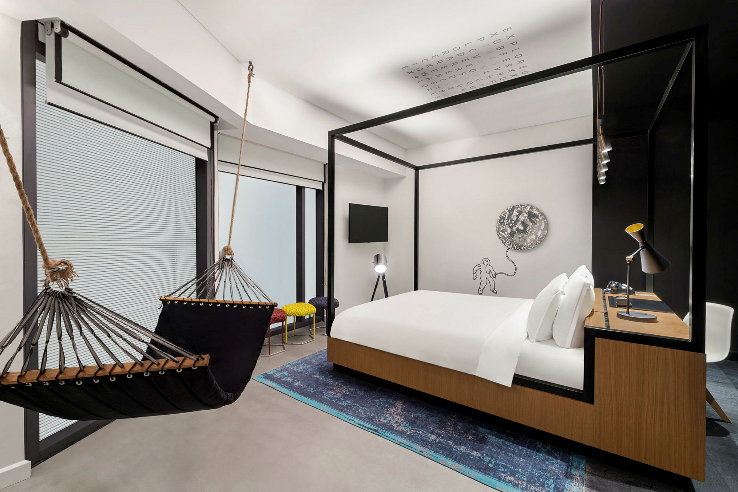 Moderni hotellihuone, jossa on riippumatto, parisänky puuelementein, työpöytä ja taiteellinen seinäkoriste. (Tekoälyn luoma teksti)