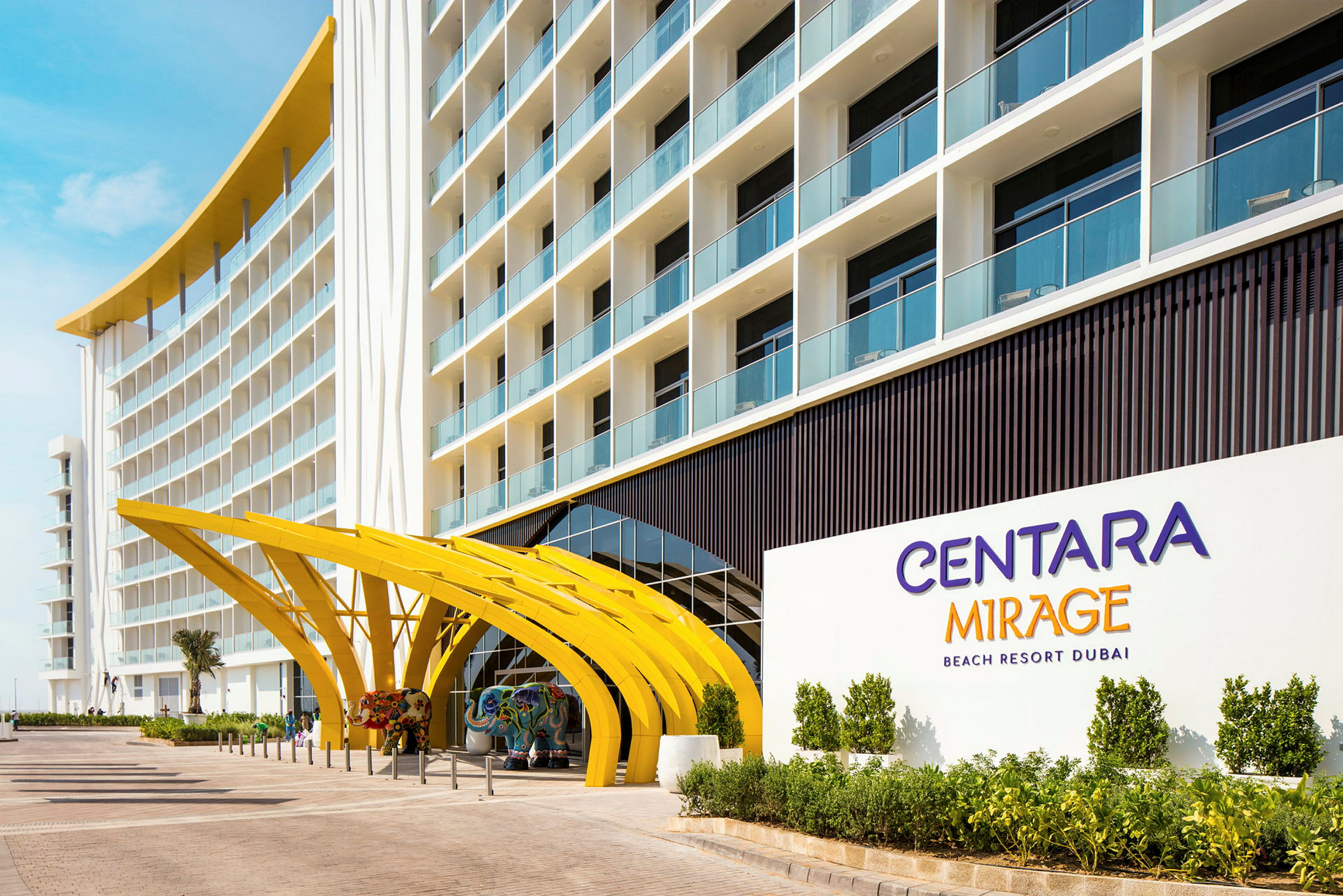 Hotellin sisäänkäynti, jossa on kaarevat keltaiset rakenteet ja kyltti, jossa lukee "Centara Mirage Beach Resort Dubai". (Tekoälyn luoma teksti)