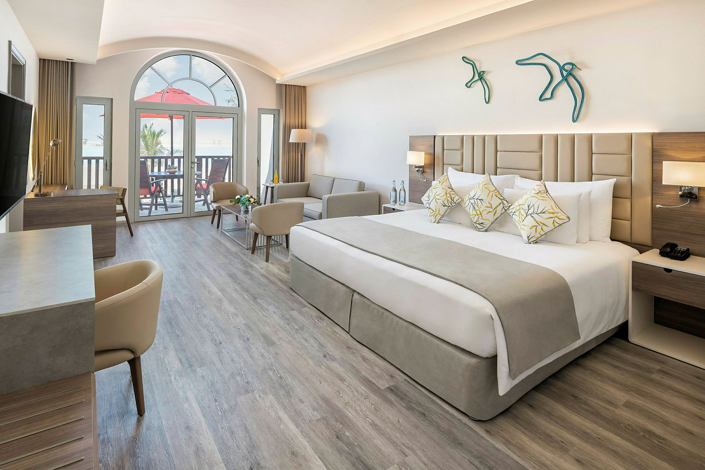 Moderni hotellihuone, jossa on leveä sänky, sohva ja työpöytä. Suuri ikkuna ja parveke taustalla. (Tekoälyn luoma teksti)