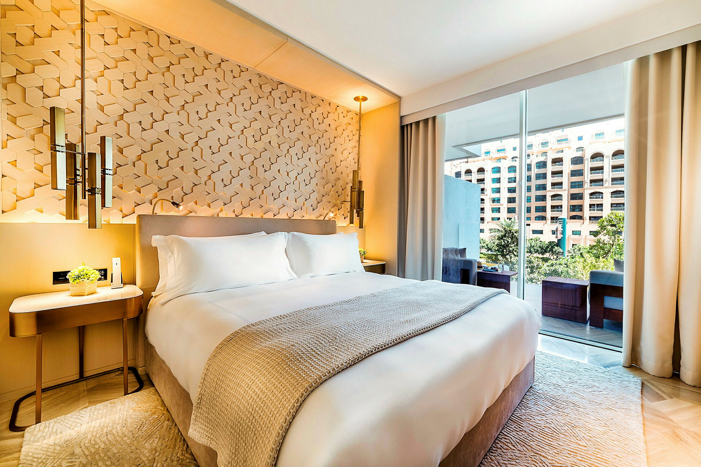 Moderni hotellihuone, jossa on valkoinen parisänky, koristeellinen seinä ja iso ikkuna, josta avautuu näkymä kerrostaloon ja vehreään ympäristöön. (Tekoälyn luoma teksti)