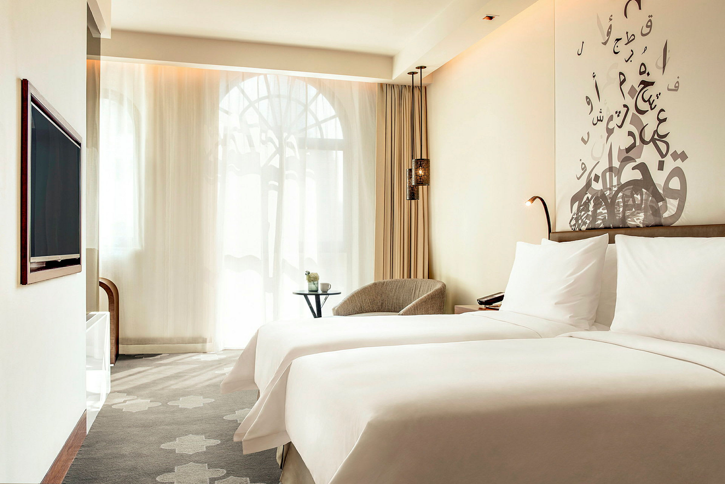 Hotellihuone, jossa on kaksi sänkyä, peitettynä valkoisilla lakanoilla. Taustalla näkyy istumapaikka, pieni pöytä ja ikkunat ohuilla verhoilla. (Tekoälyn luoma teksti)