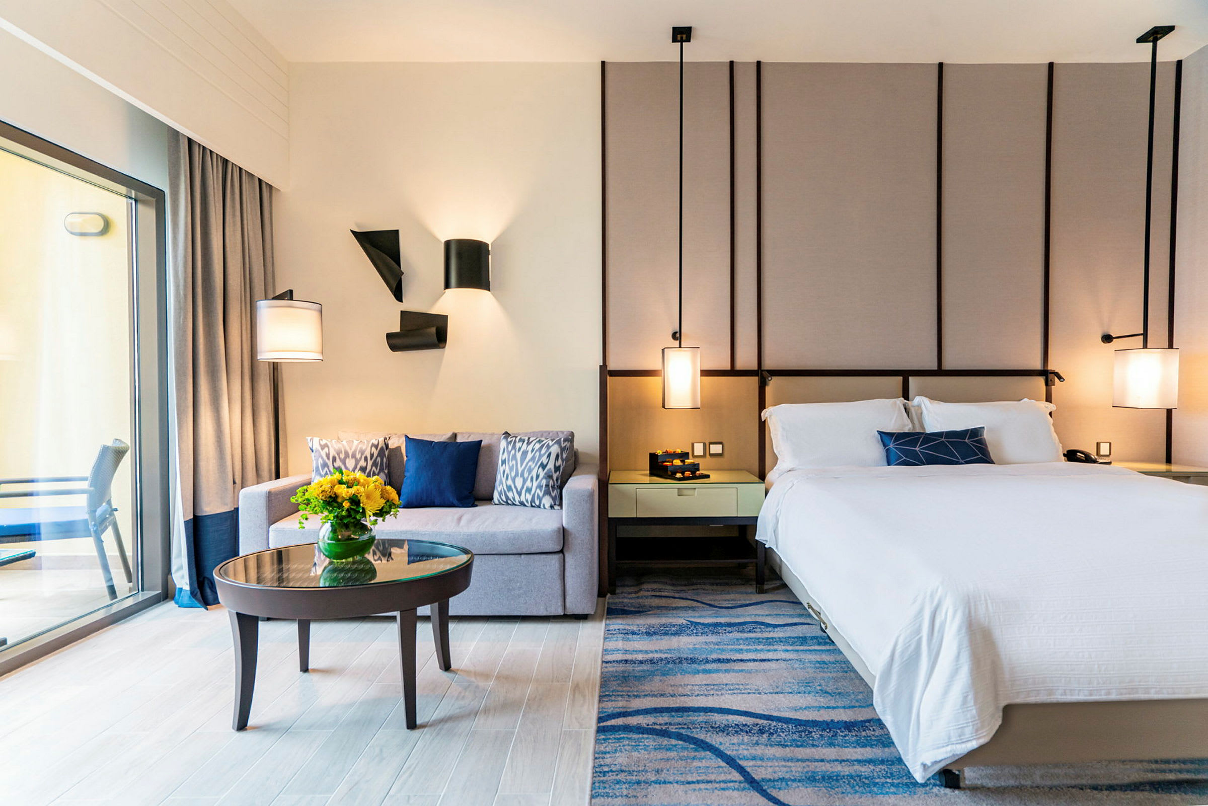 Moderni hotellihuone, jossa on sänky, sohva, pieni sohvapöytä kukkavaasilla sekä parveke, joka näkyy lasioven kautta. (Tekoälyn luoma teksti)