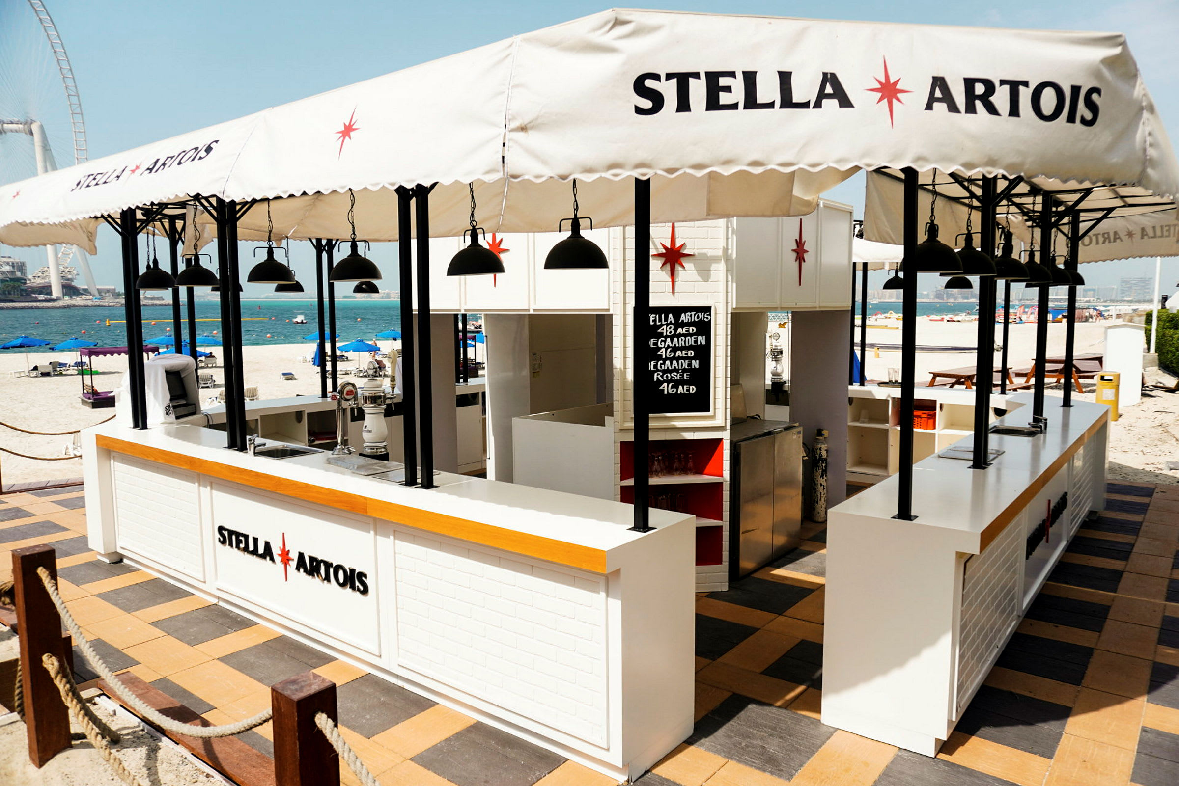 Rantabaari ja Stella Artois -logo, aurinkovarjoja ja pöytiä. (Tekoälyn luoma teksti)