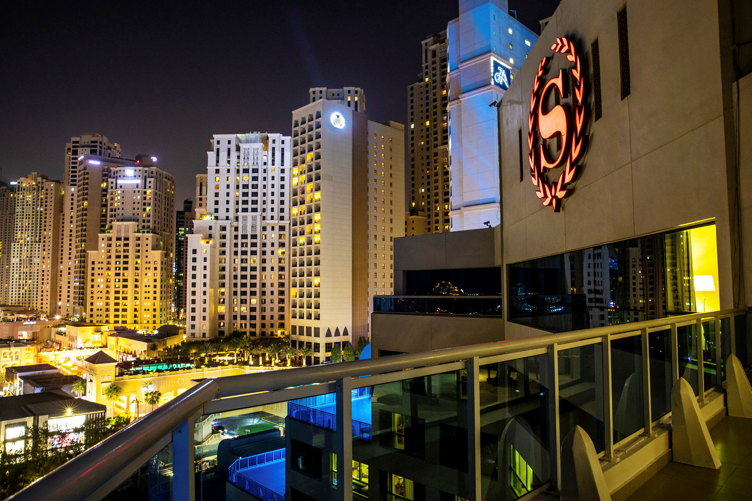 Yökuva kaupungista, jossa on valaistuja korkeiden rakennusten julkisivuja sekä etualalla rakennus, jossa on logotunnus. (Tekoälyn luoma teksti)