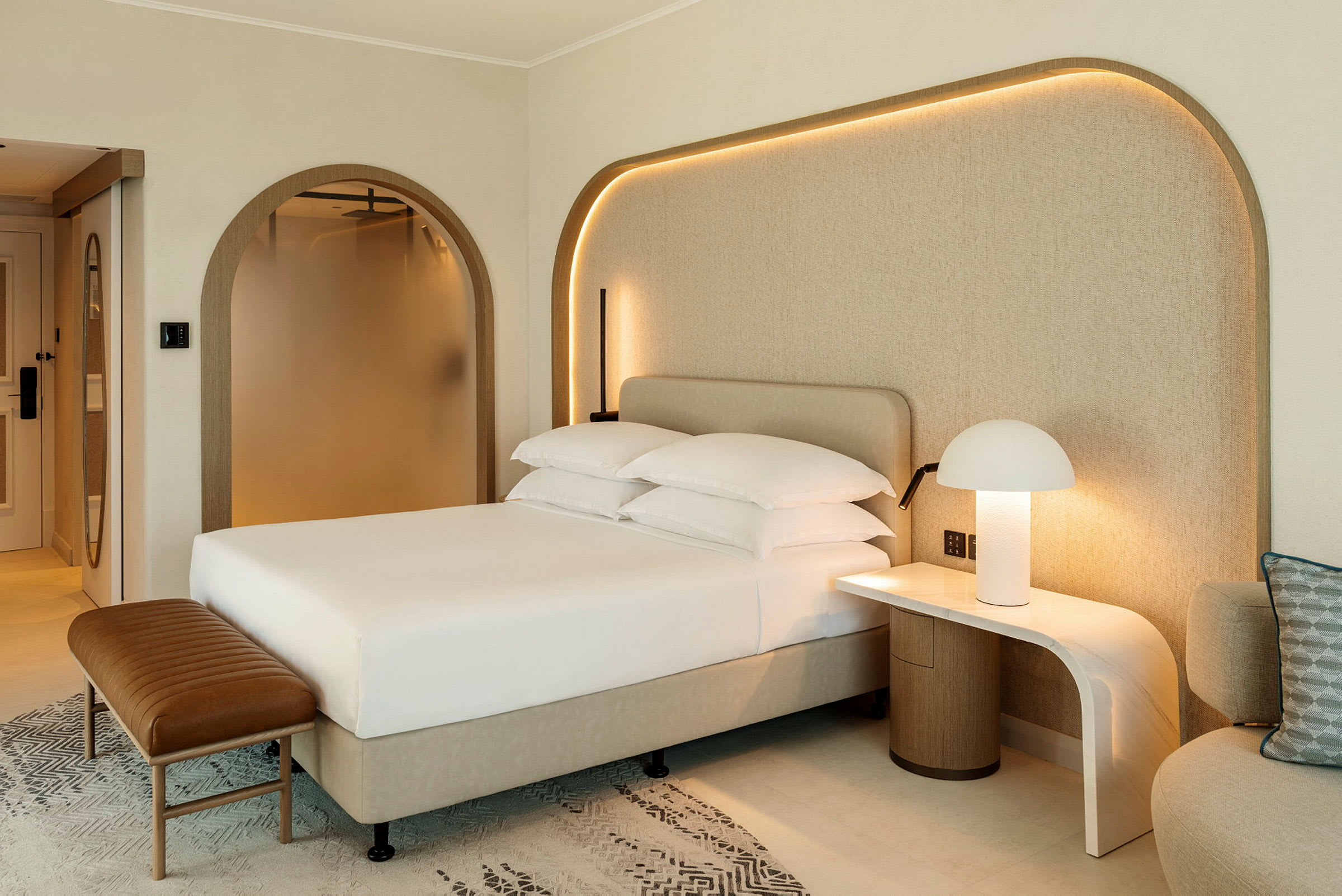 Kuva modernista hotellihuoneesta, jossa on pedattu sänky, yöpöytä lampulla ja istuin. (Tekoälyn luoma teksti)