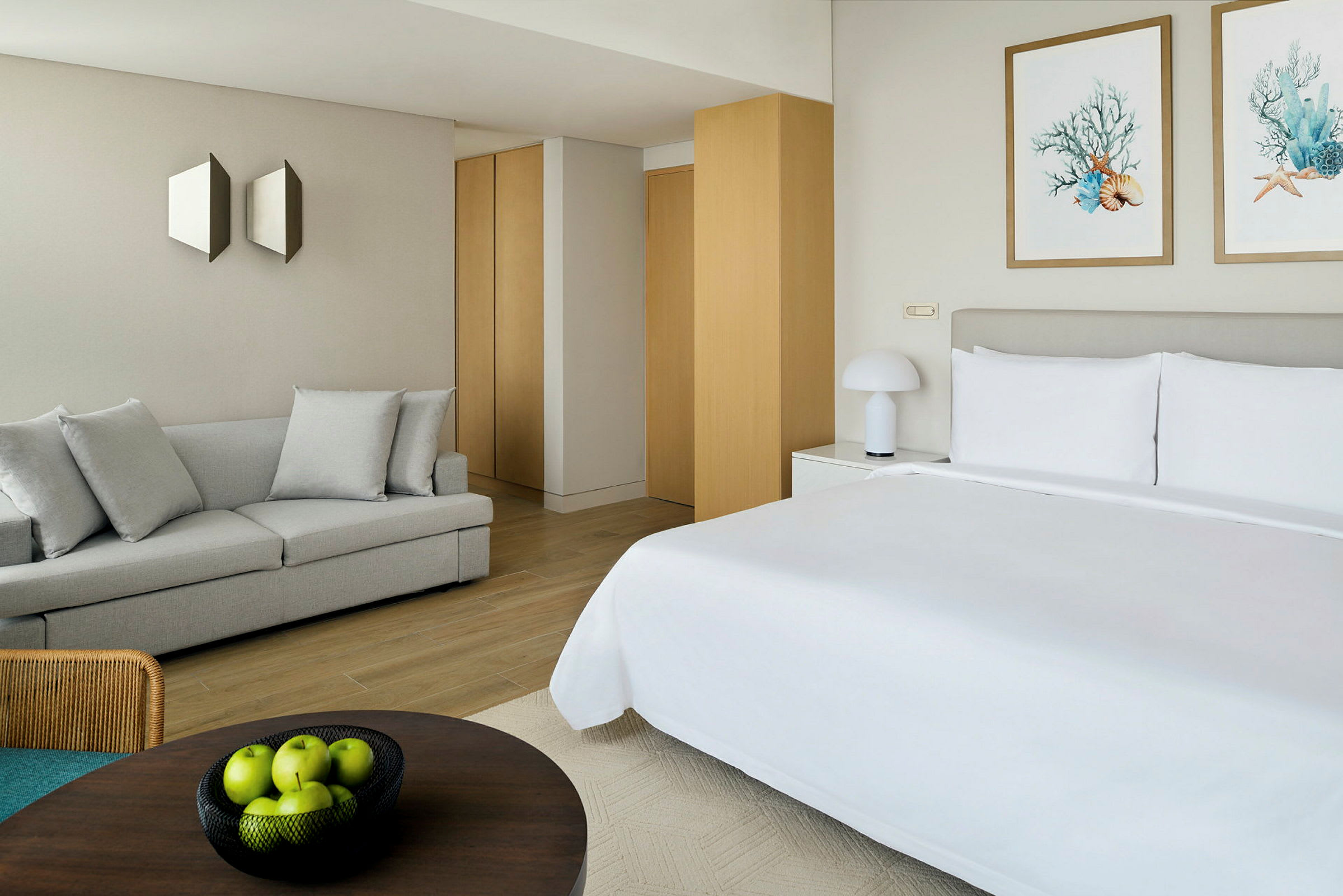 Moderni hotellihuone, jossa on pedattu parisänky. Huoneessa on myös vaalea sohva, seinäkoristeita ja pöydällä oleva kulho vihreitä omenoita. (Tekoälyn luoma teksti)