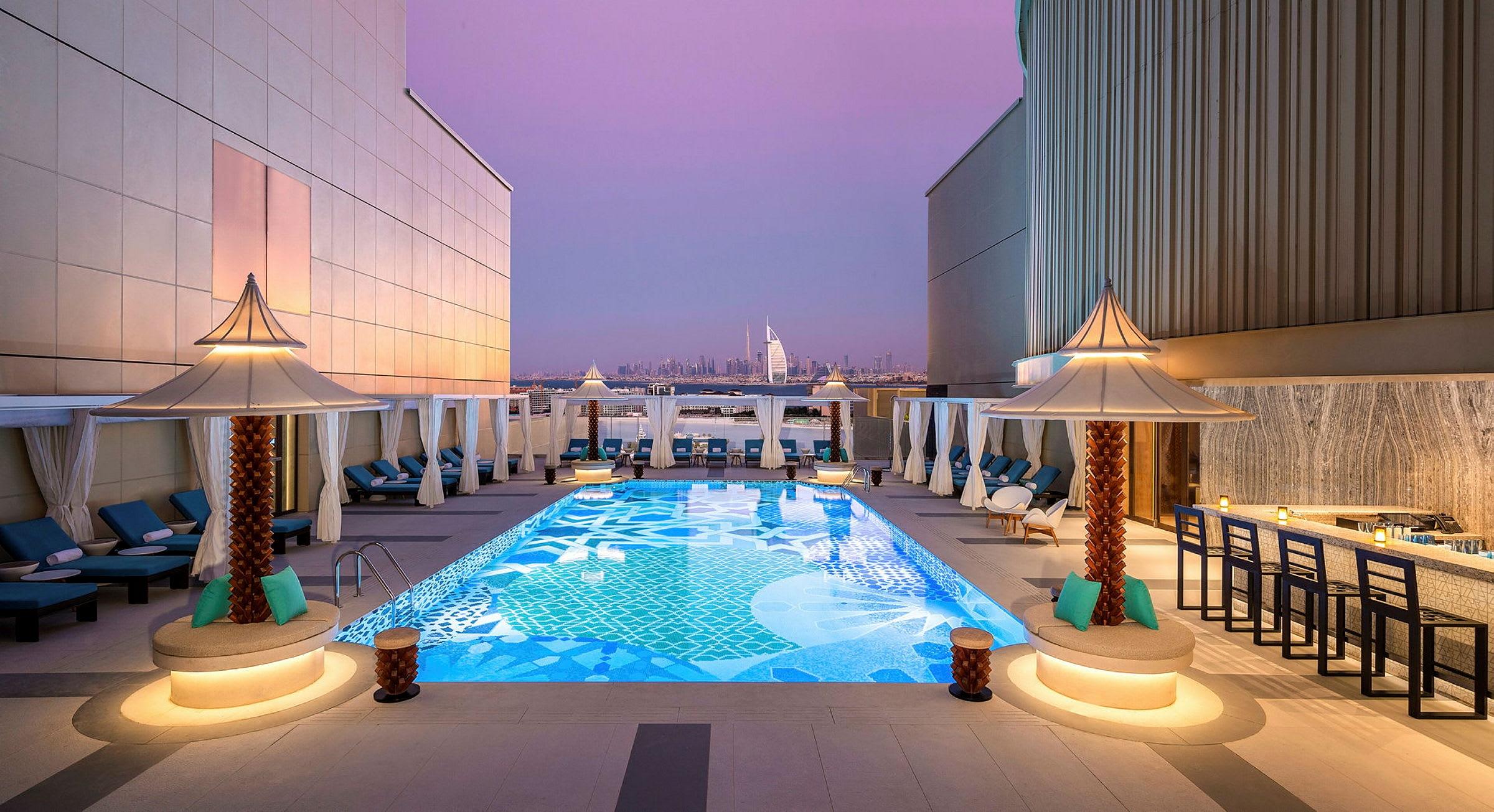Kattoterassi, jossa koristeellinen uima-allas, jota ympäröivät aurinkotuolit ja pienet aurinkovarjot. Taustalla näkyy Burj Al Arab ja Dubain kaupunkimaisema violetin iltataivaan alla. (Tekoälyn luoma teksti)