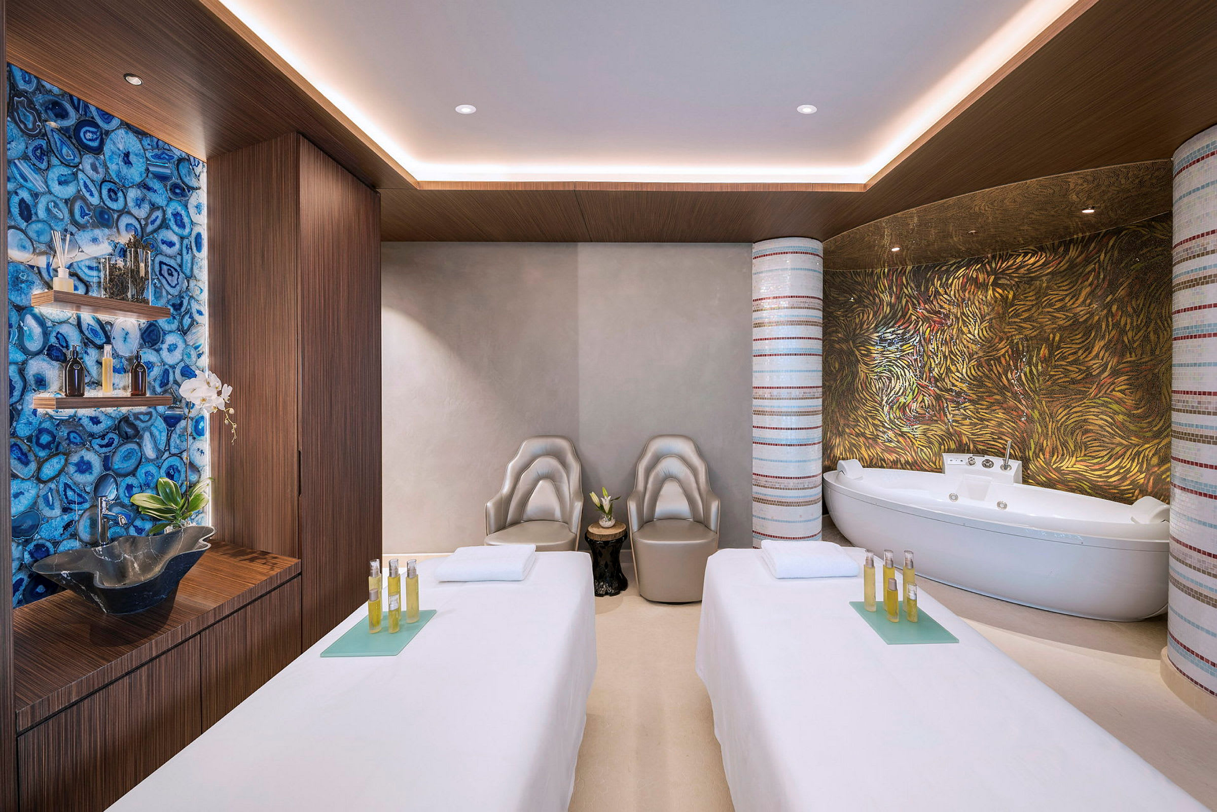 Spa-huone, jossa on kaksi hierontapöytää, modernit tuolit ja kylpyamme. Taustalla värikkäitä kuvioituja seiniä ja hyllyjä koristeilla. (Tekoälyn luoma teksti)