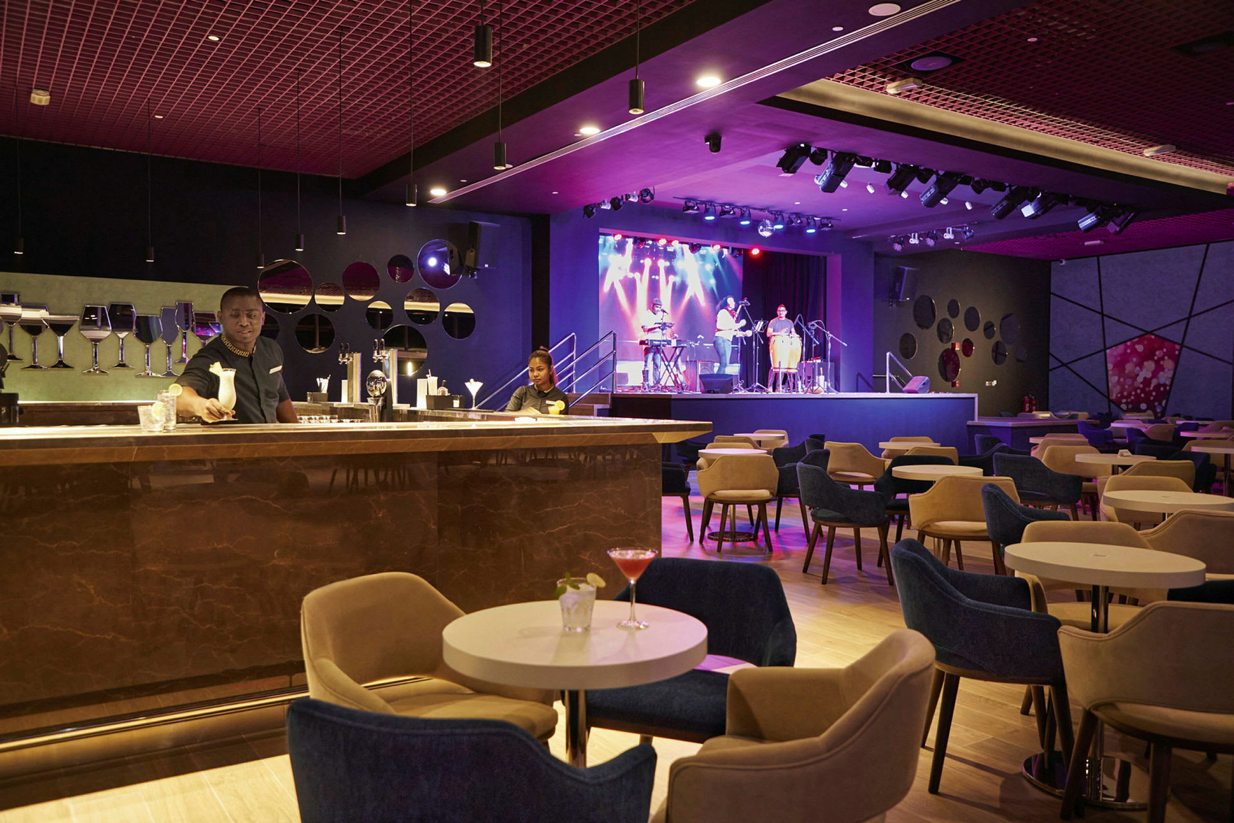 Tyylikäs lounge, jossa istumapaikoja pienien pöytien ympärillä, etualalla baaritiski ja taustalla lava muusikoineen. (Tekoälyn luoma teksti)