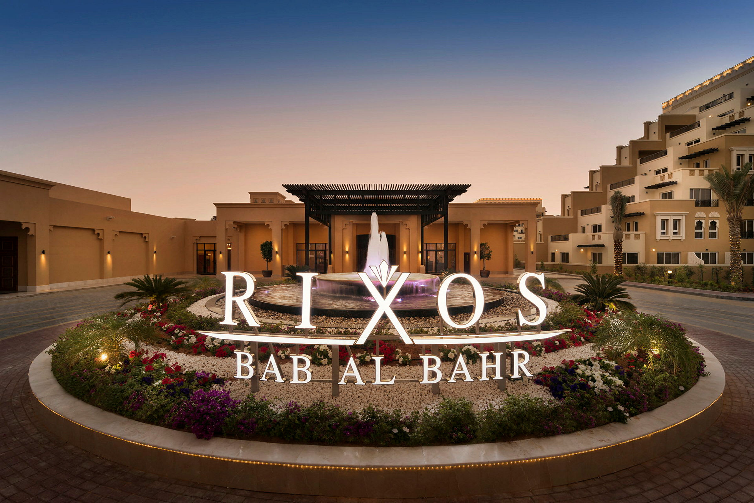 Hotellin sisäänkäynti, suihkulähde ja ympäröiviä kukkia, kyltti tekstillä Rixos Bab Al Bahr. (Tekoälyn luoma teksti)