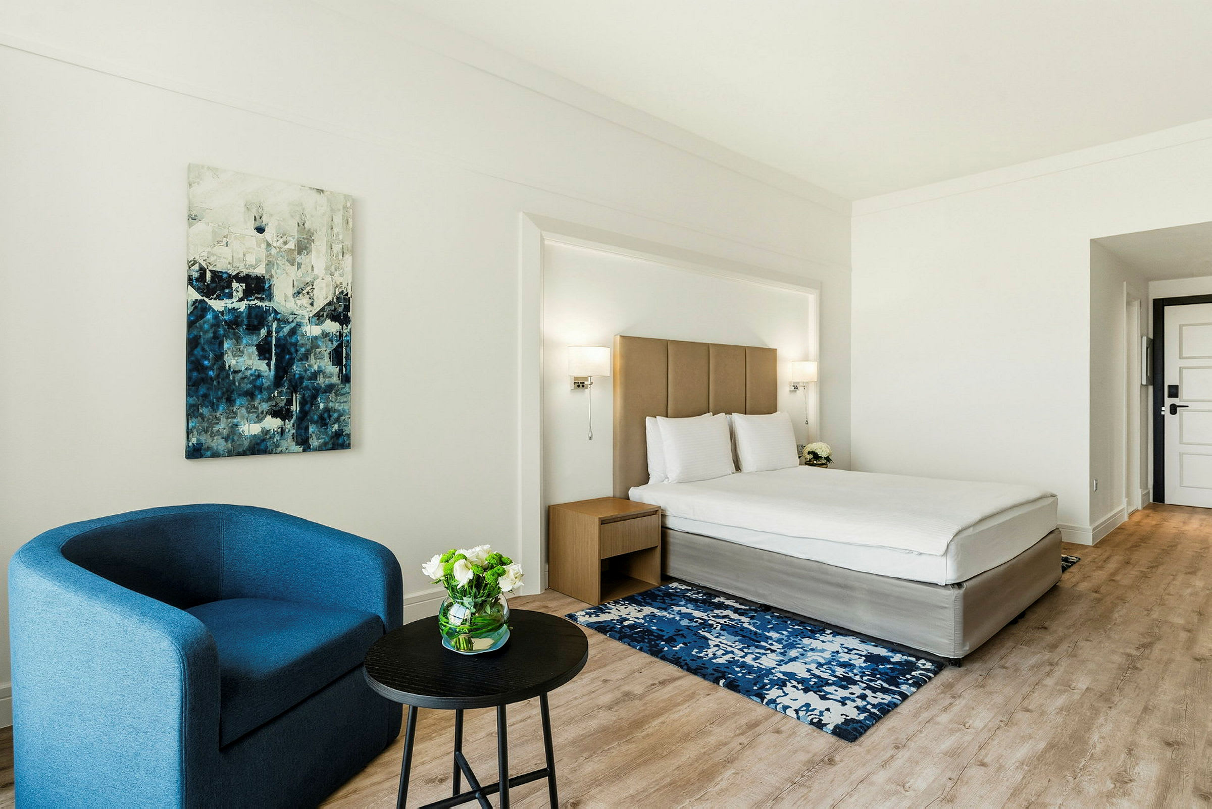 Hotellihuone, jossa on sijattu sänky, sininen nojatuoli, sohvapöytä ja maalaus seinällä. (Tekoälyn luoma teksti)