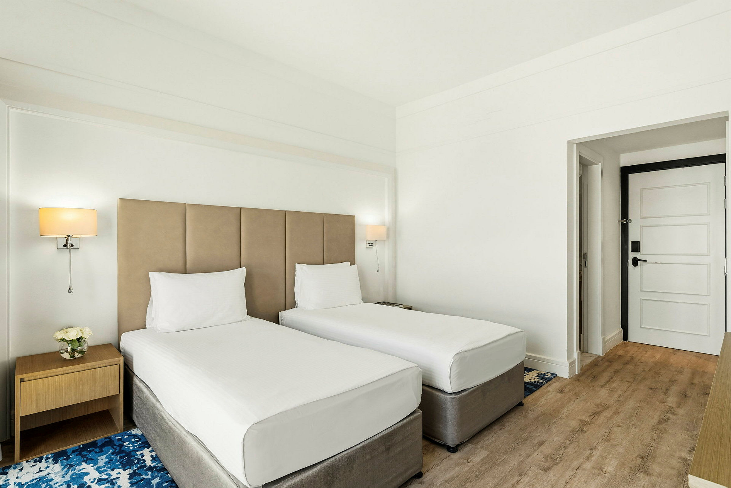 Hotellihuone, jossa on kaksi yhden hengen sänkyä, yöpöytä ja ovi. (Tekoälyn luoma teksti)