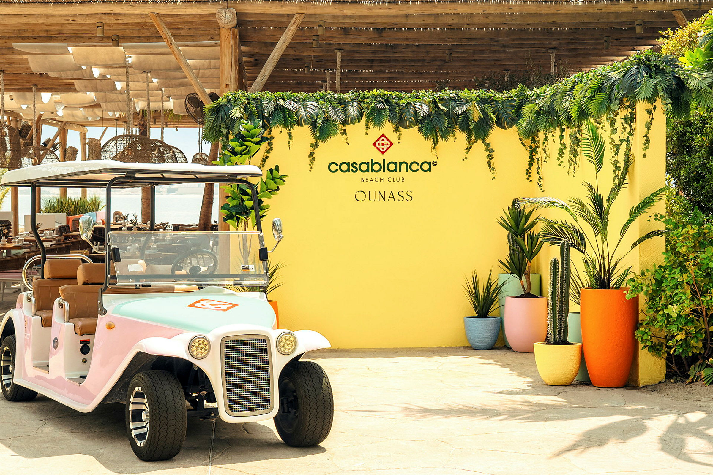 Golf-auton edessä on keltainen seinä, joka on koristeltu kasvillisuudella ja kukilla sekä tekstillä 'Casablanca Beach Club'. (Tekoälyn luoma teksti)