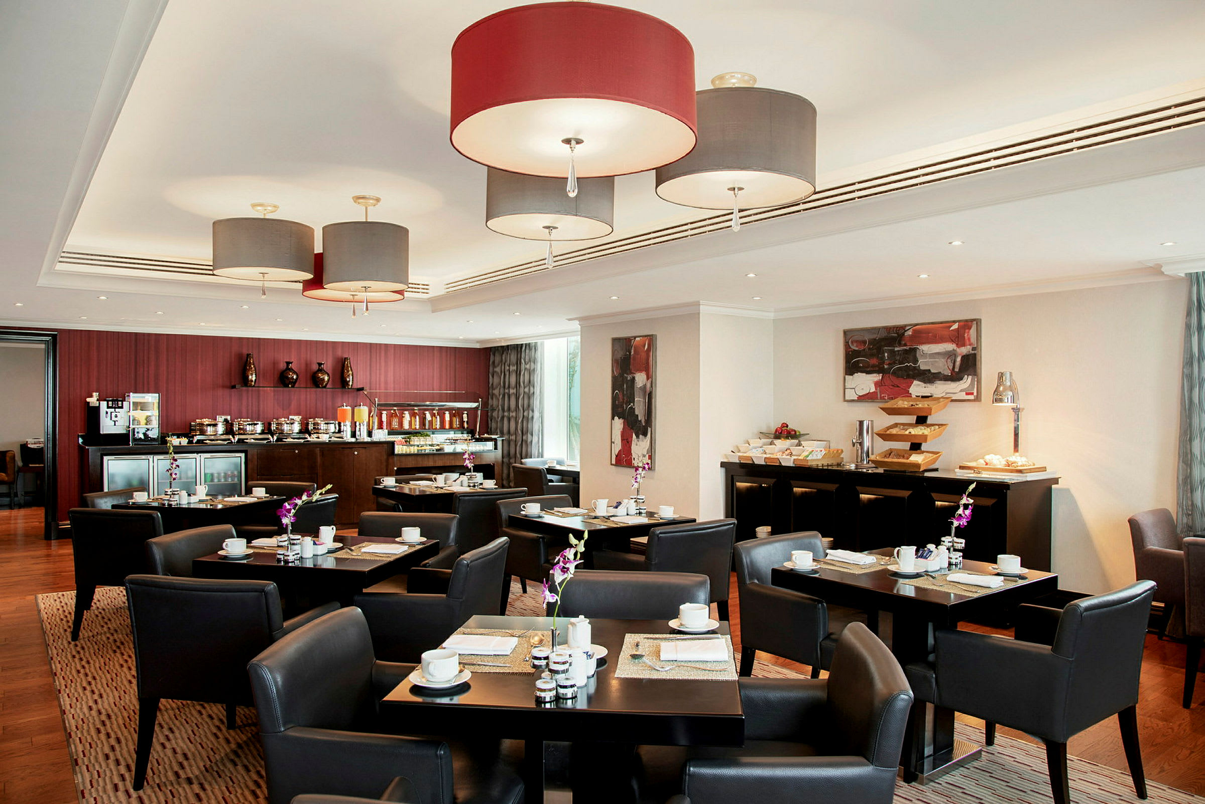 Bistroalue, jossa on pöytiä ja tuoleja buffetpöydän ympäröimänä. Katosta riippuvat valaisimet ja seinillä oleva taide täydentävät sisustuksen. (Tekoälyn luoma teksti)
