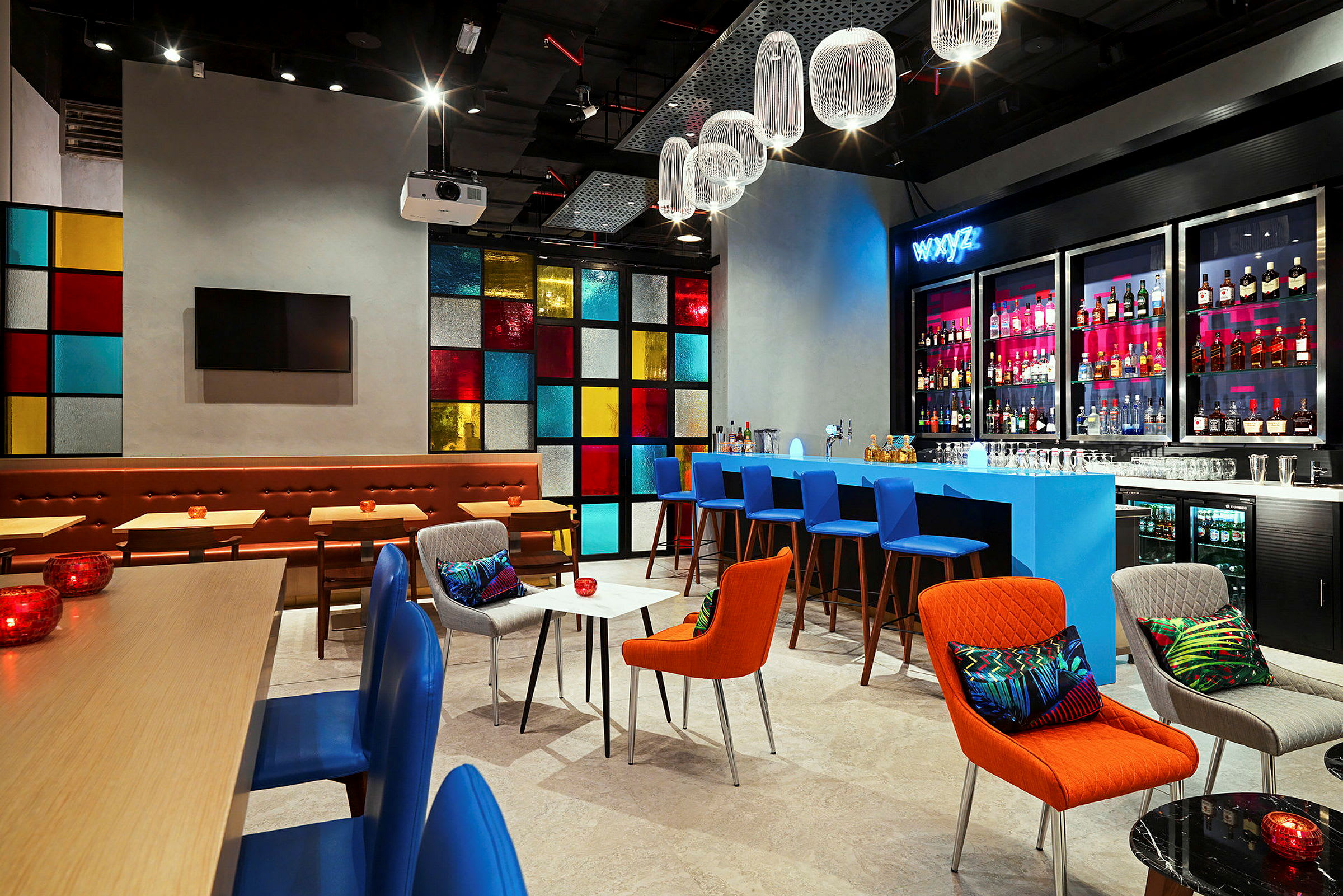 Moderni baari tai lounge, jossa on värikäs sisustus, sininen baaritiski ja hyllyjä pullotaustalla. Erivärisistä ruuduista koostuva ikkunapaneeli luo eloisan taustan. Valaistus on lämmin, ja suunnittelu on elegantti ja tyylikäs. (Tekoälyn luoma teksti)