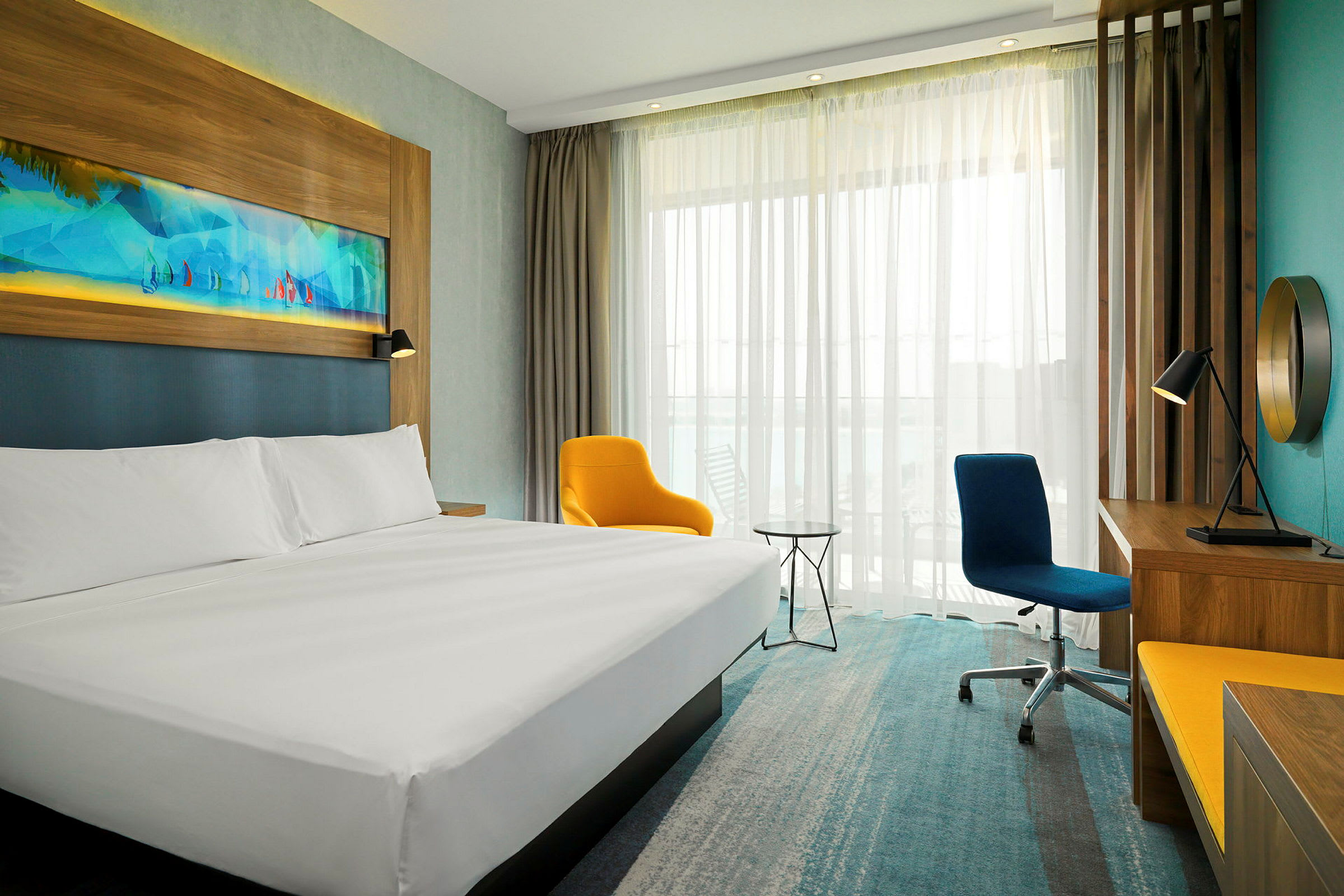 Moderni hotellihuone, jossa on suuri sänky, keltainen nojatuoli, kirjoituspöytä ja peili. Panoraamaikkuna vaaleilla verhoilla näkyy taustalla. (Tekoälyn luoma teksti)