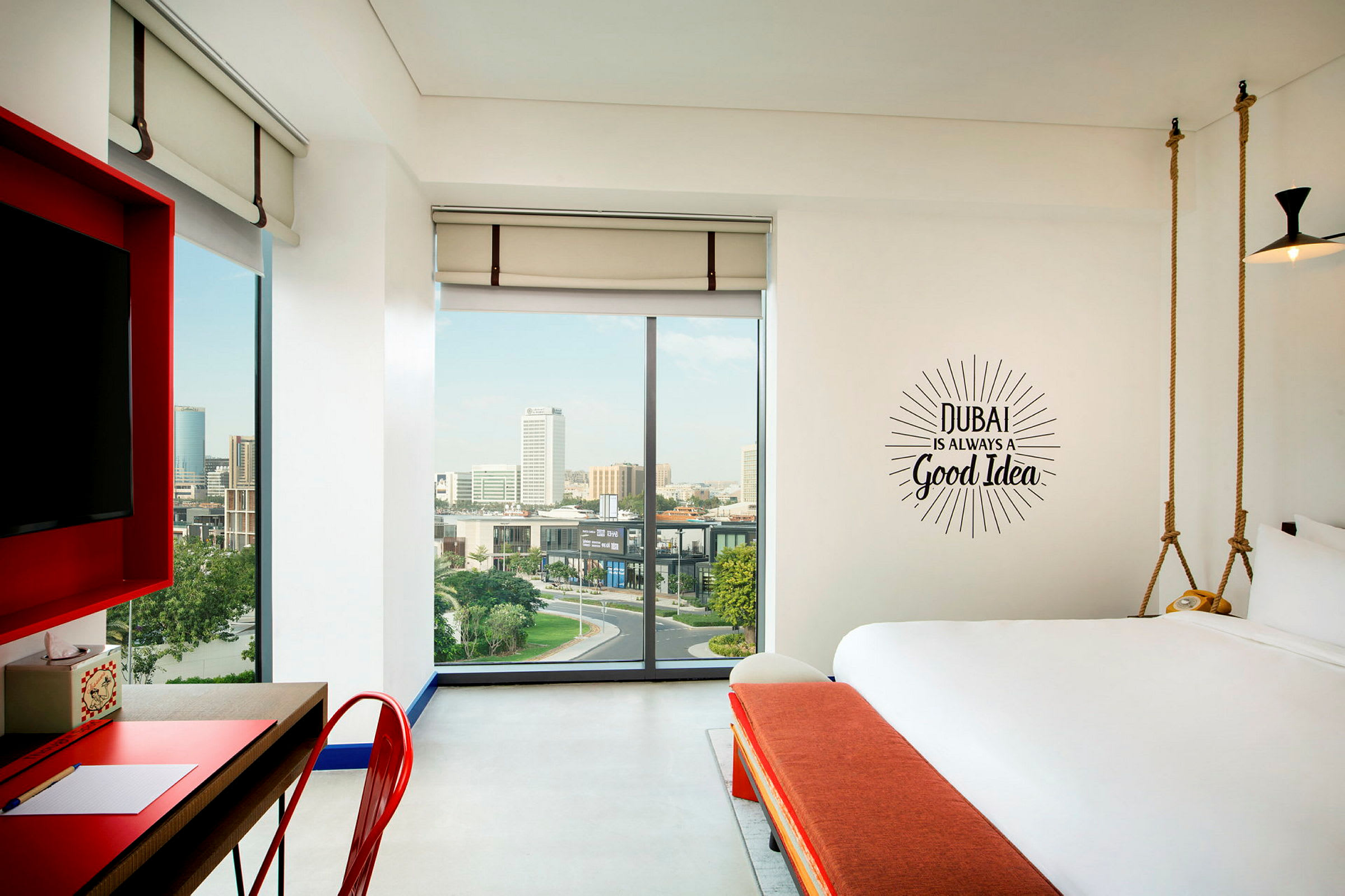 Moderni hotellihuone suurilla ikkunoilla ja näkymällä kaupunkiin. Seinällä lukee teksti: "Dubai is always a good idea". (Tekoälyn luoma teksti)