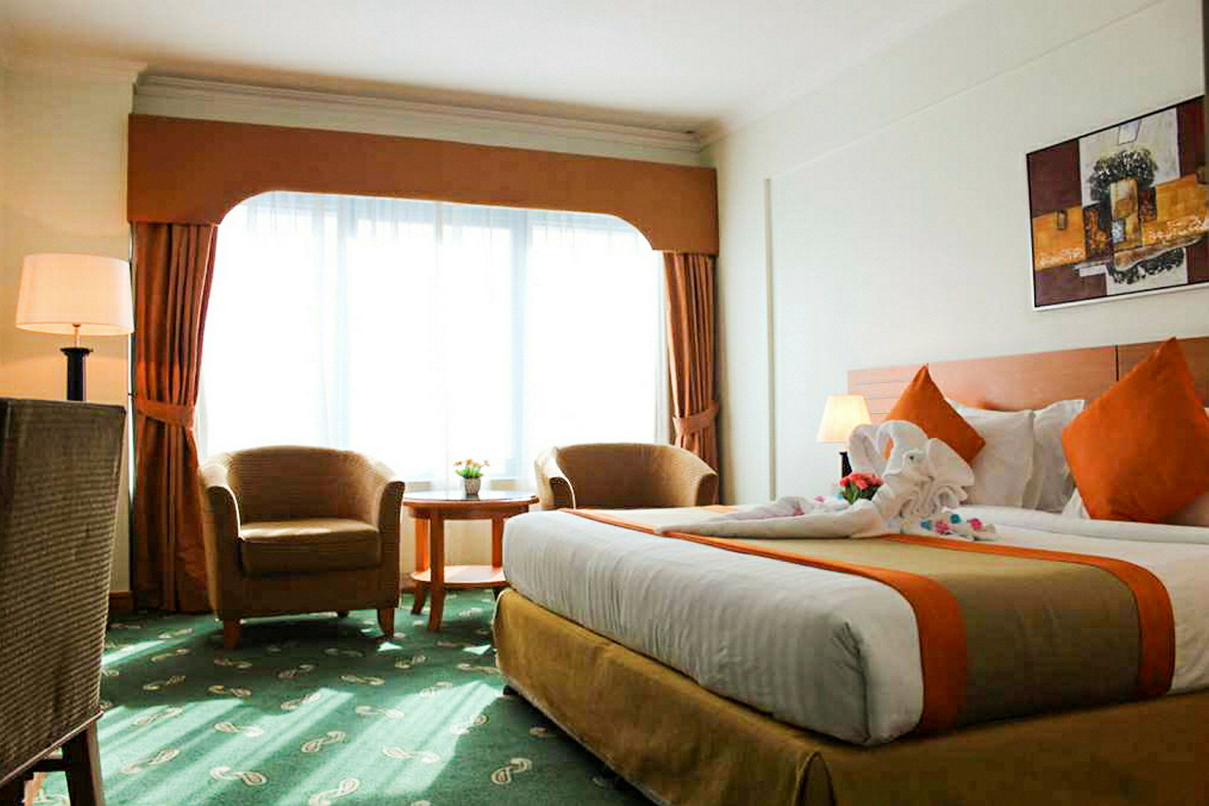 Hotellihuone, jossa on suuri sänky, kaksi nojatuolia, pyöreä pöytä ja suuret verhoilla varustetut ikkunat. (Tekoälyn luoma teksti)