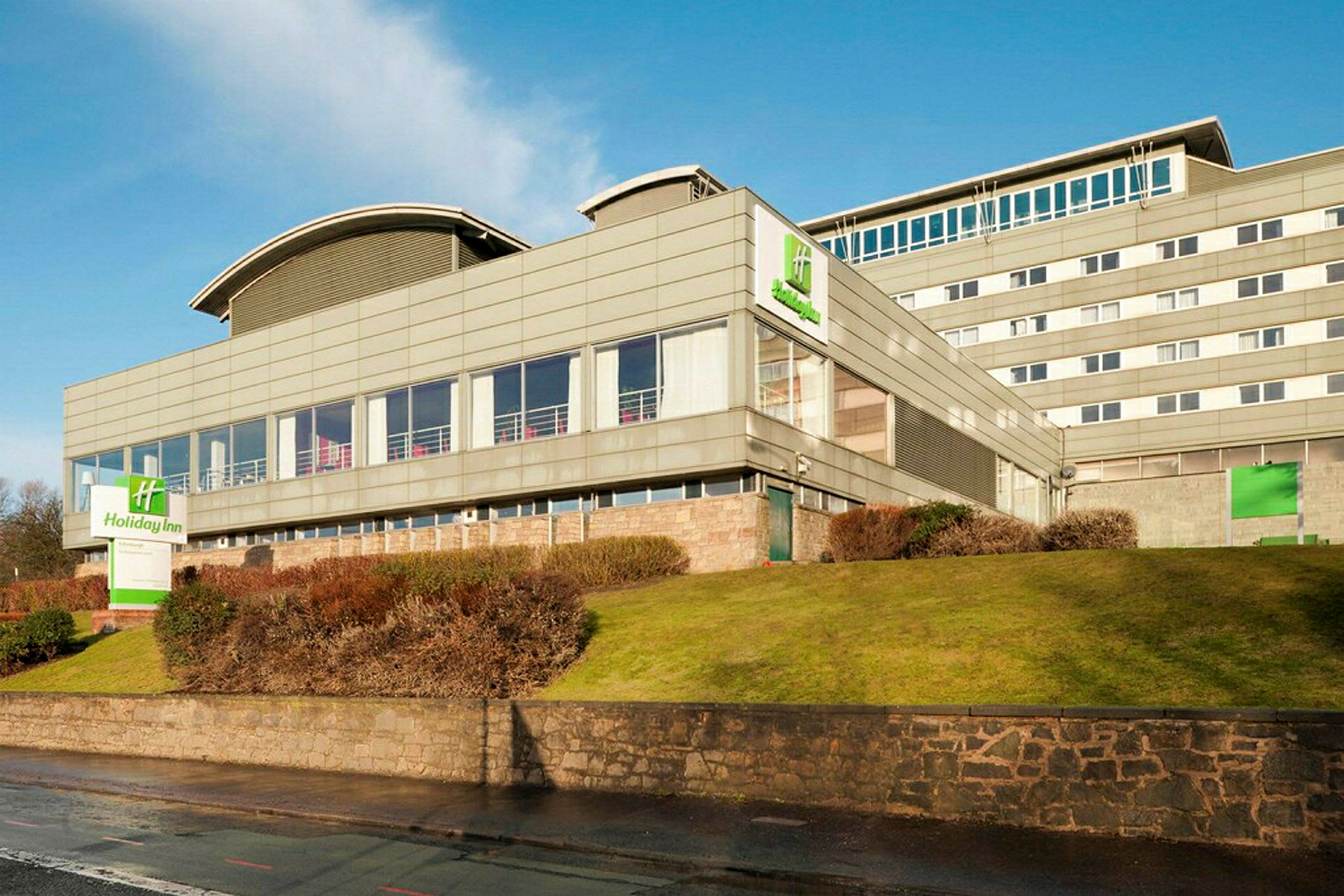 Holiday Inn -hotelli modernissa rakennuksessa, jota ympäröivät nurmikko ja pensaat. (Tekoälyn luoma teksti)