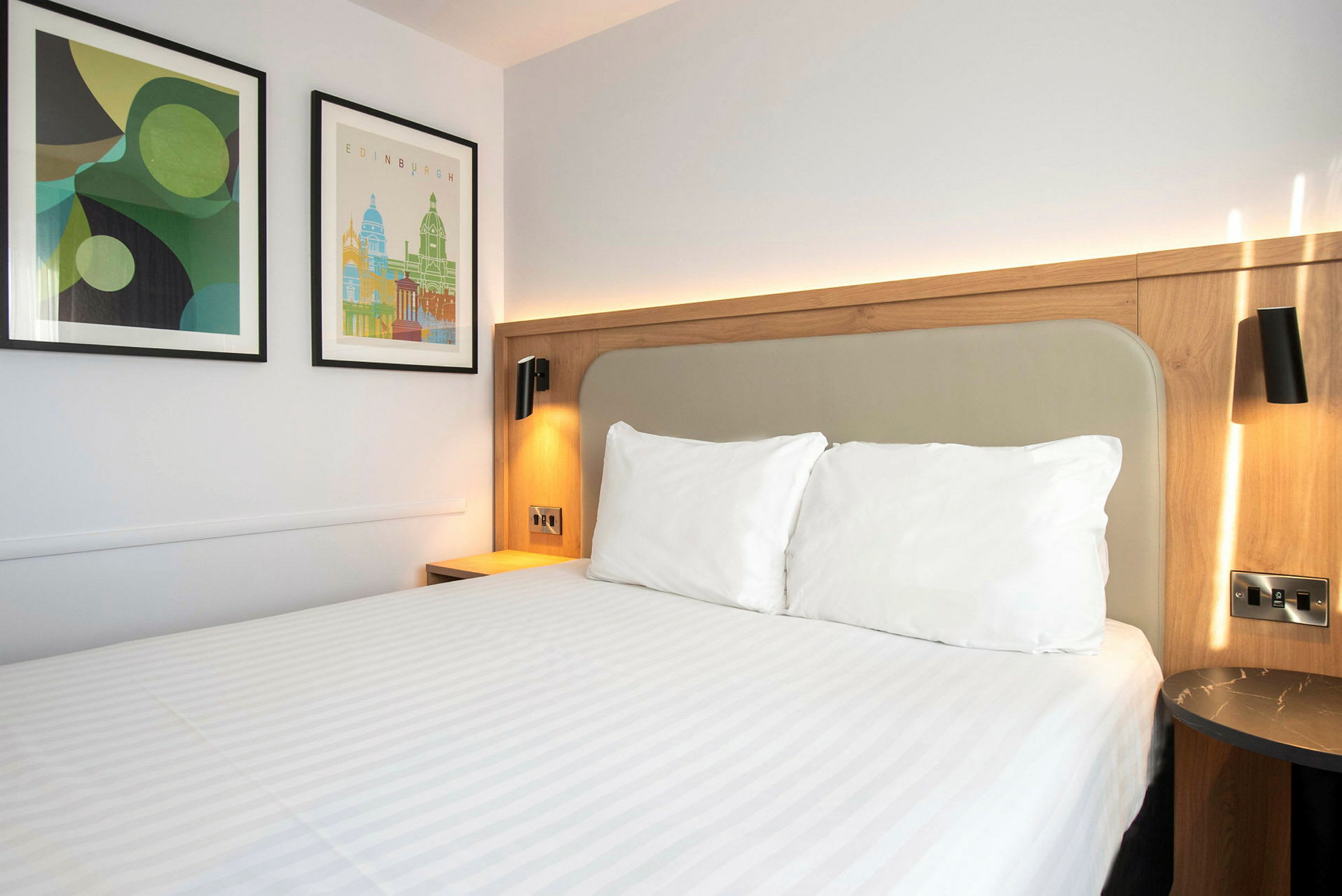 Hotellihuoneen sänky, jossa on valkoiset vuodevaatteet ja kaksi taulua seinällä. (Tekoälyn luoma teksti)