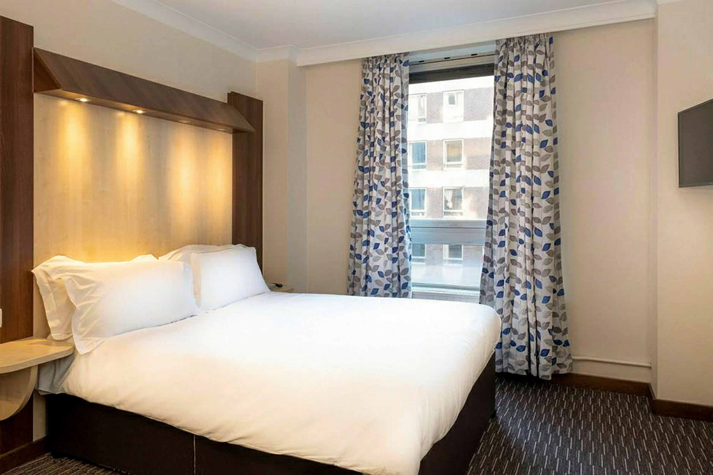 Hotellihuone, jossa on parivuode, ikkuna kuviollisilla verhoilla ja seinään kiinnitetty televisio. (Tekoälyn luoma teksti)
