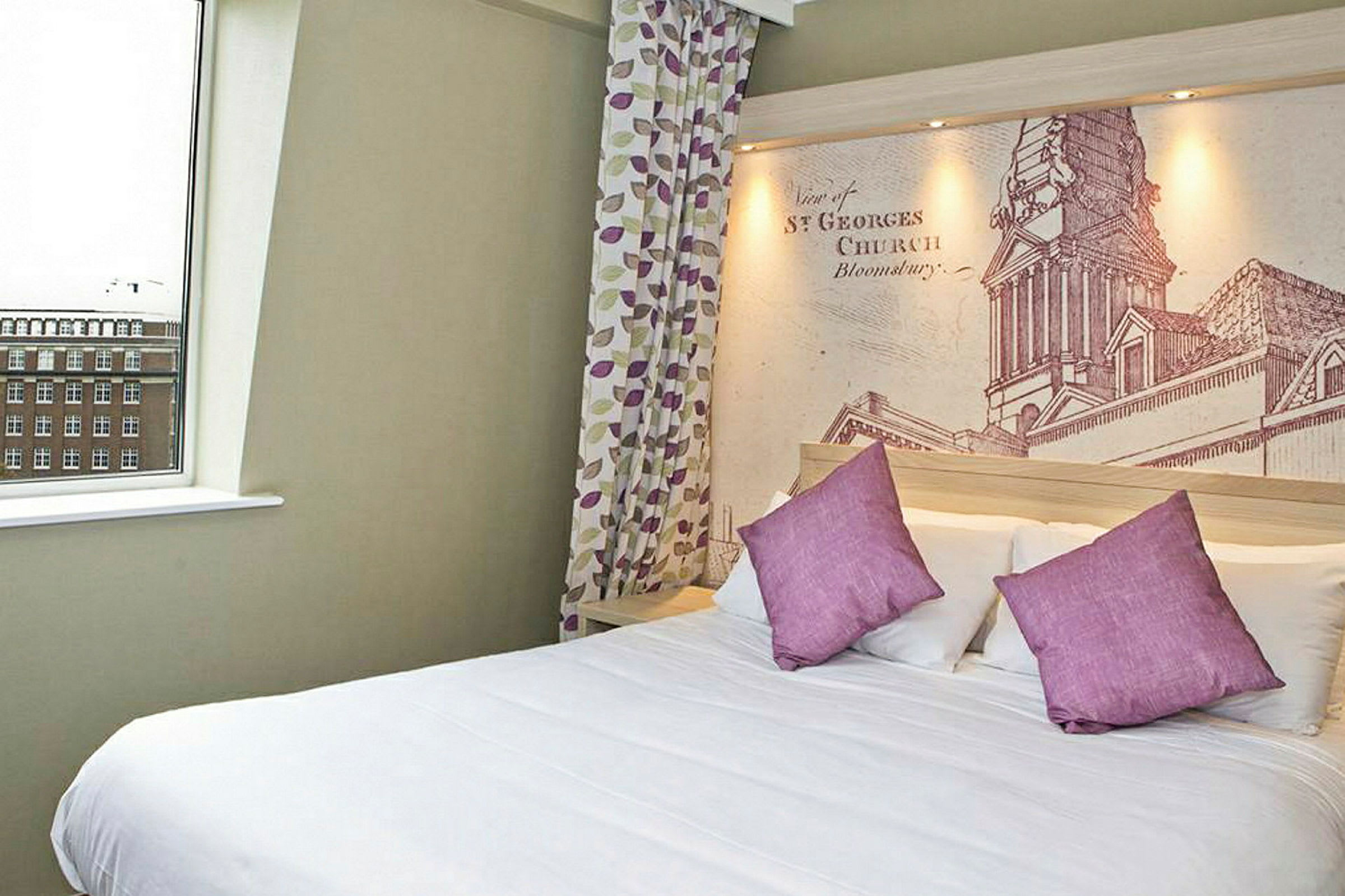 Hotellihuone, jossa suuri sänky, violetit tyynyt ja yksityiskohtainen seinämaalaus sängyn takana. (Tekoälyn luoma teksti)