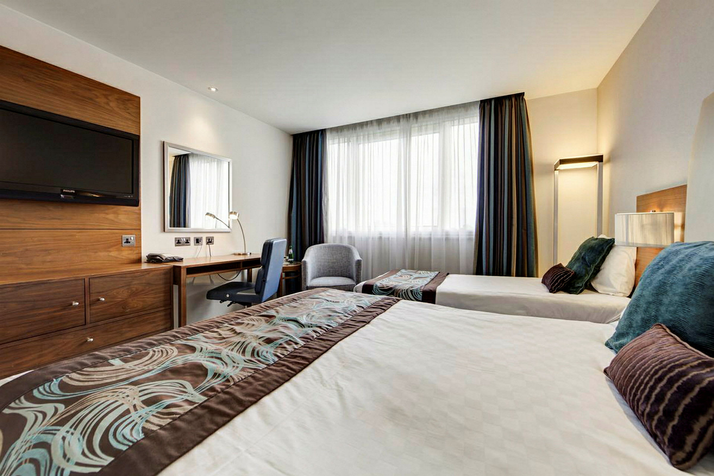Hotellihuone, jossa on kaksi yhden hengen vuodetta, kirjoituspöytä ja seinällä televisio. (Tekoälyn luoma teksti)