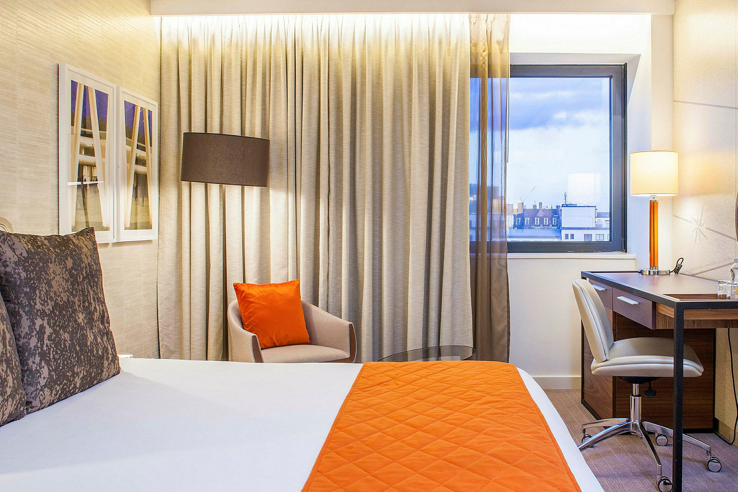 Hotellihuone, jossa on oranssi päiväpeite ja tyyny, tuoli ja kirjoituspöytä ikkunan lähellä. (Tekoälyn luoma teksti)