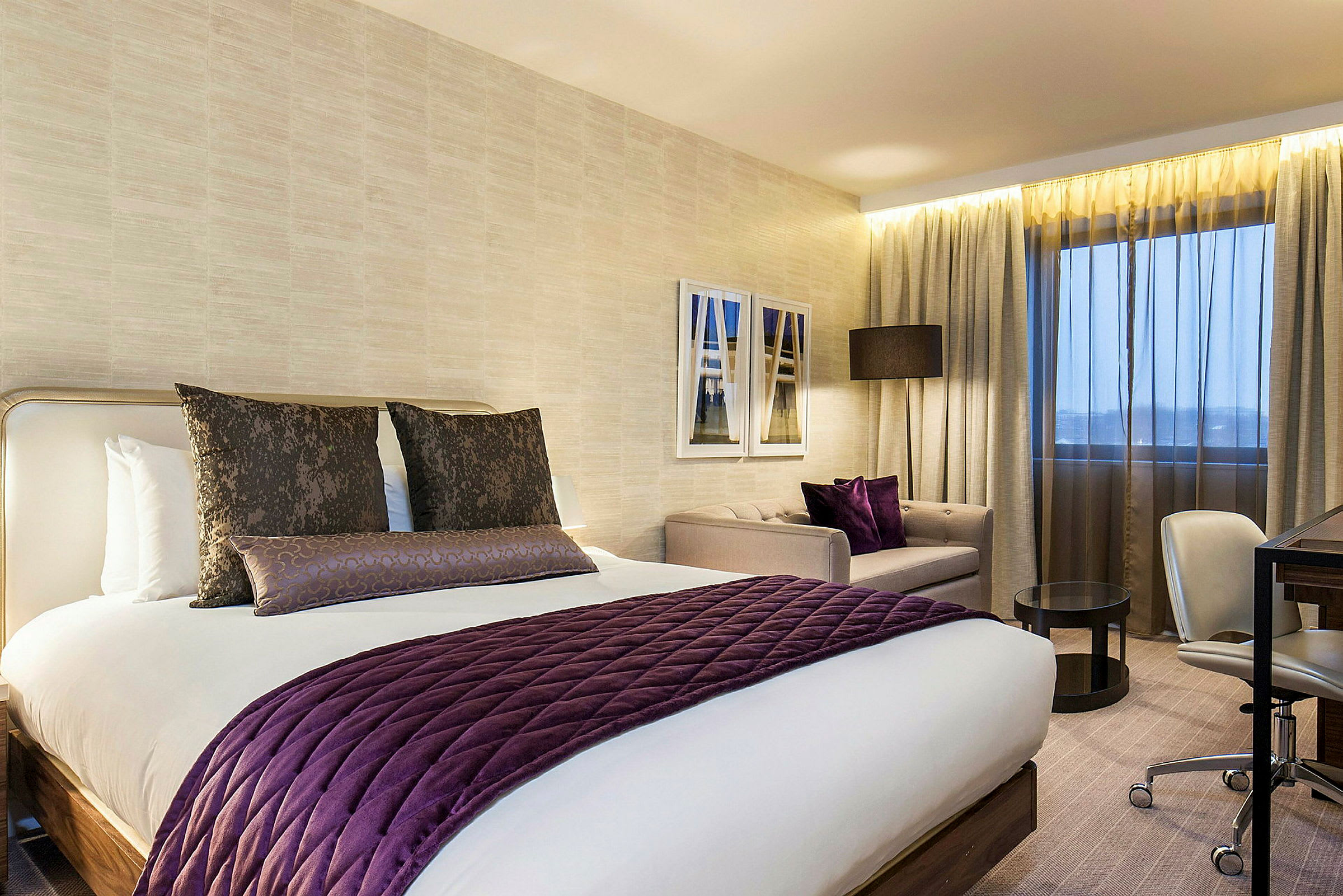Modernisti sisustettu hotellihuone, joka sisältää suuren sängyn violetilla yksityiskohdilla, istuinryhmän ja työpöydän. (Tekoälyn luoma teksti)