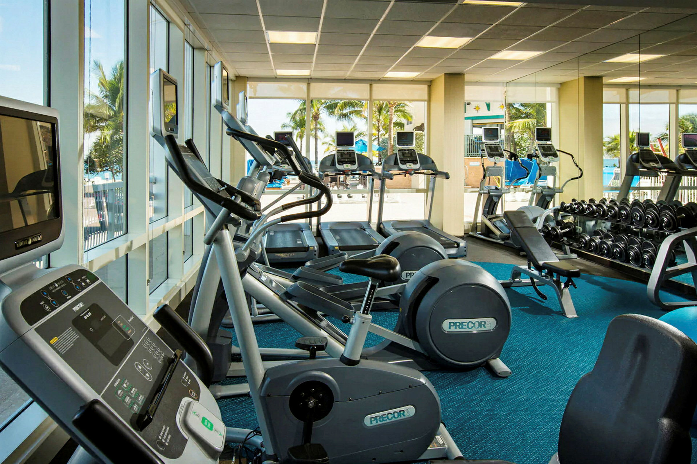 Kuntosali, jossa on kuntolaitteita ja irtopainoja, suuret ikkunat tarjoavat näkymän ulos. (Tekoälyn luoma teksti)