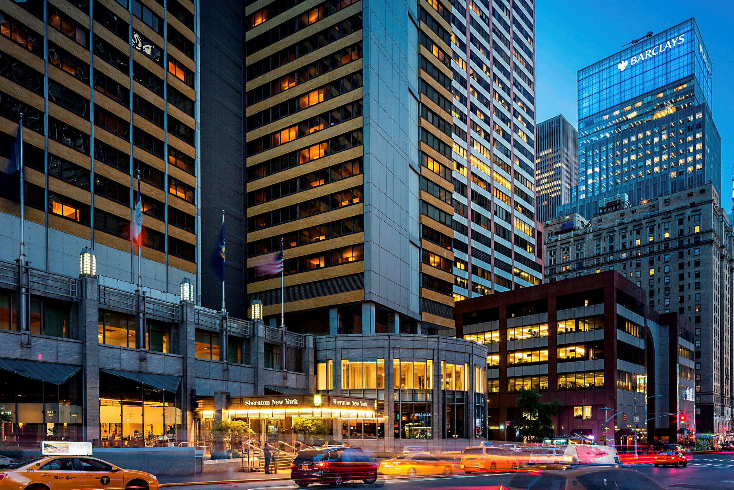 Kuva kaupunkimaisemasta, jossa on korkeita rakennuksia. Yhdessä rakennuksessa lukee "Sheraton New York" sen sisäänkäynnillä. Takseja näkyy kadulla. (Tekoälyn luoma teksti)