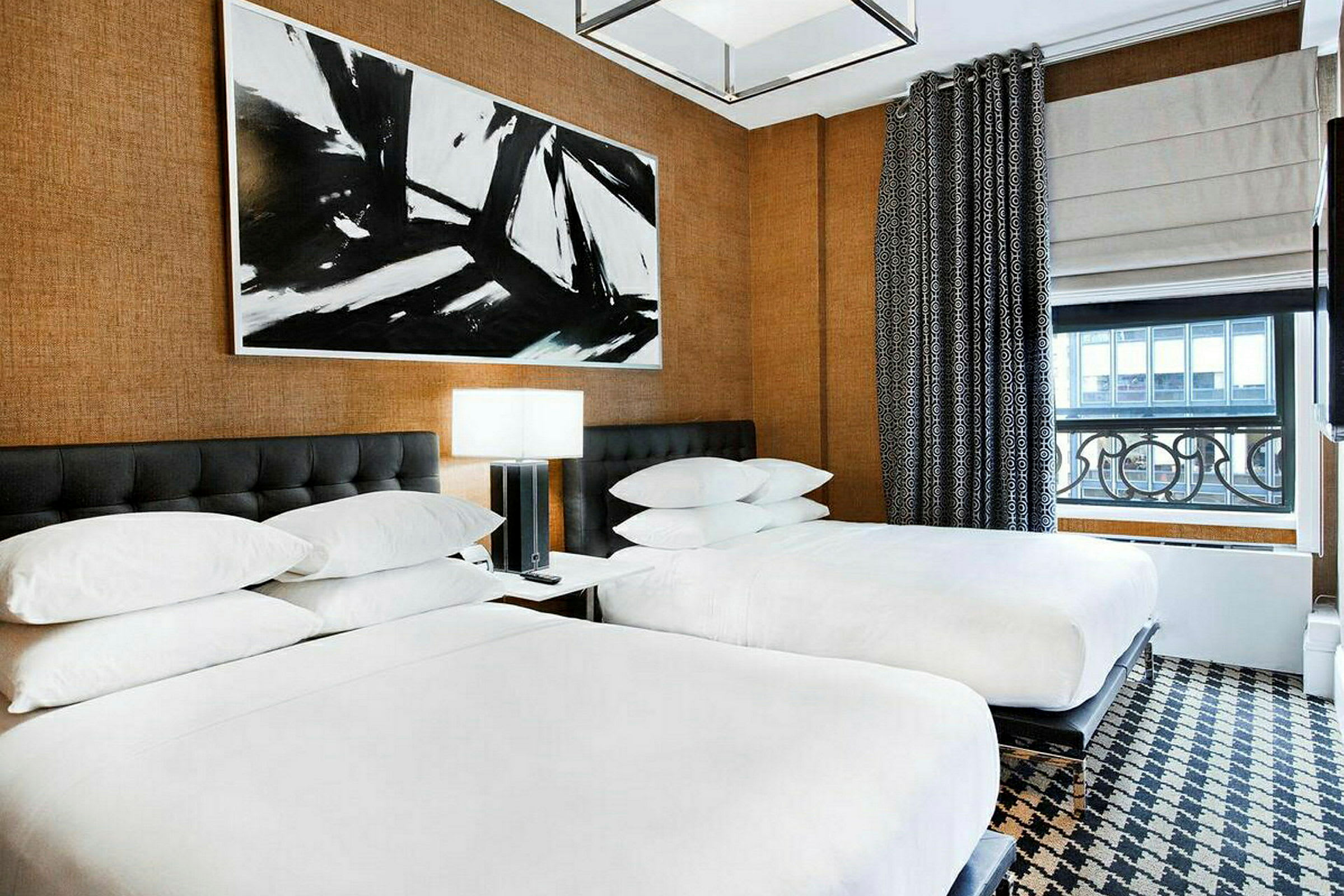 Hotellihuone, jossa on kaksi erillistä vuodetta, yksinkertaiset yöpöydät lampuilla ja suuri abstrakti maalattu taulu sänkyjen yläpuolella. (Tekoälyn luoma teksti)
