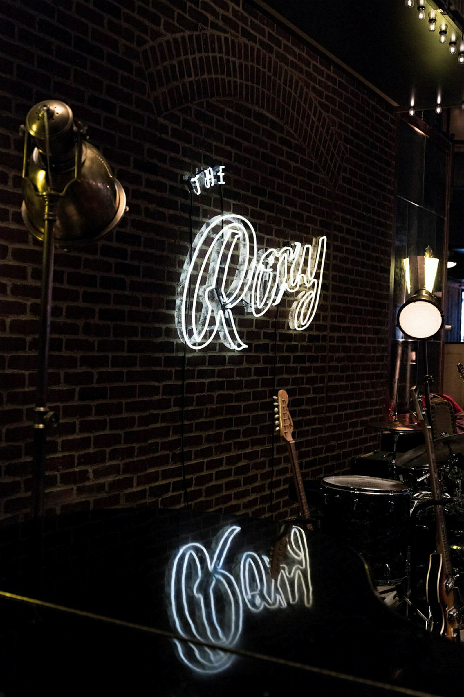 Seinä, jossa on neonlamppu, joka kirjoittaa "The Roxy" valkoisilla kirjaimilla, sekä taustalla näkyy soittimia, kuten kitara ja rummut. (Tekoälyn luoma teksti)