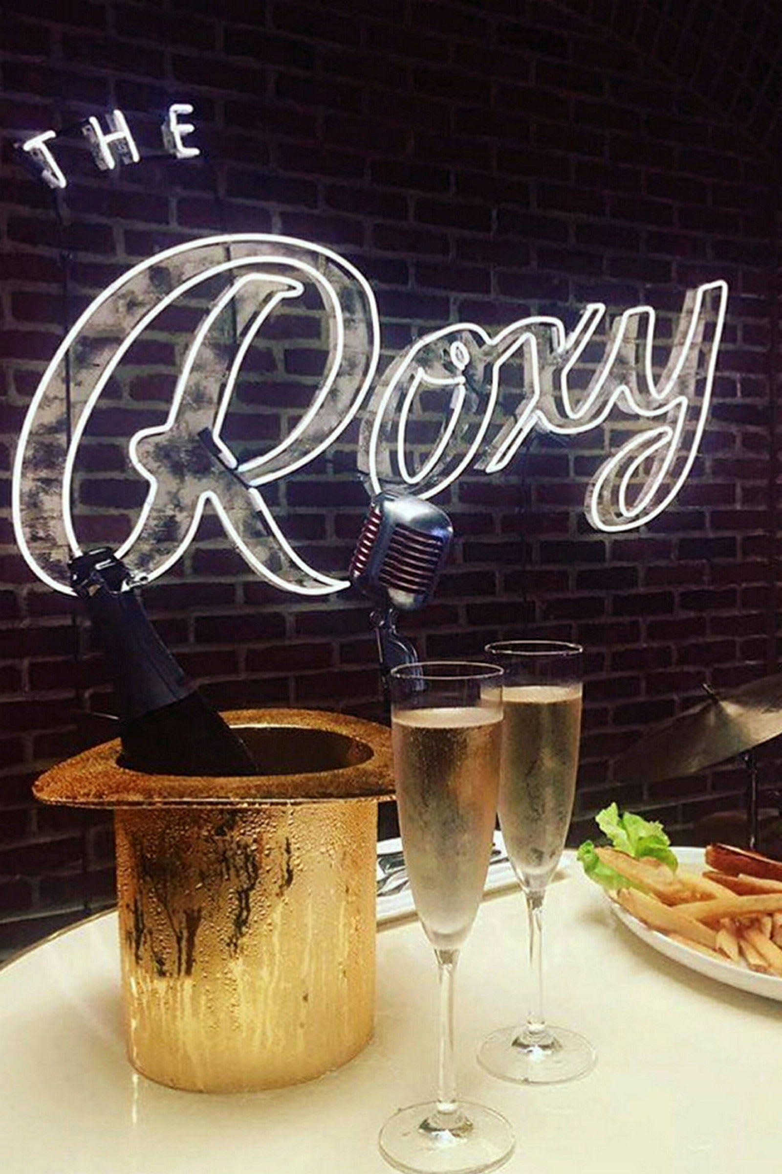 Pöytä, jolla kaksi lasia kuohuviiniä, samppanjapullo ämpärissä, joka on hatun muodossa, sekä lautanen ranskalaisia. Taustalla näkyy neonteksti "The Roxy". (Tekoälyn luoma teksti)