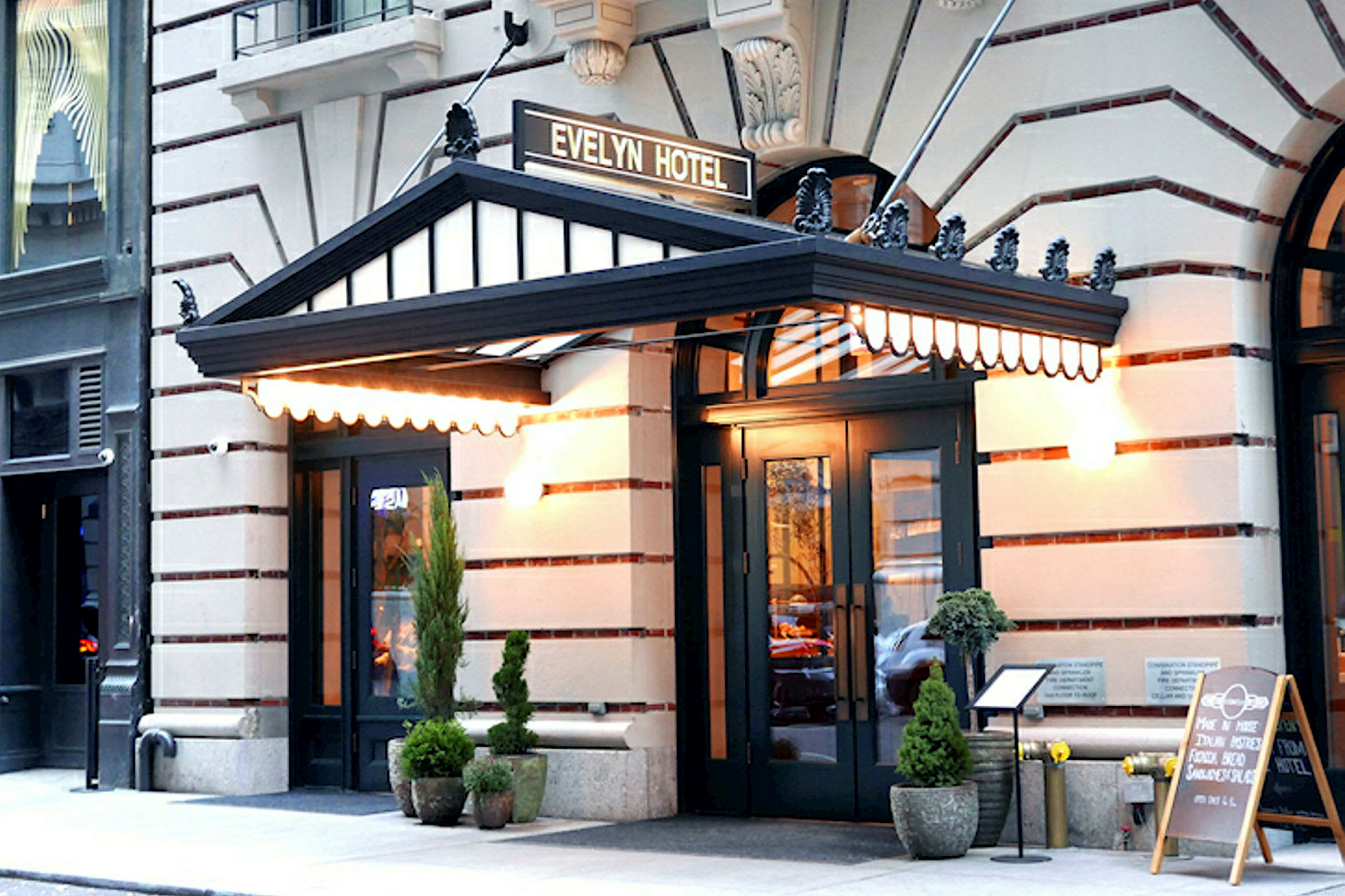 Hotellin sisäänkäynti, jossa on musta markiisi ja "Evelyn Hotel" -kyltti yläpuolella. Ulkona on ruukkukasveja ja menu jalustalla. (Tekoälyn luoma teksti)