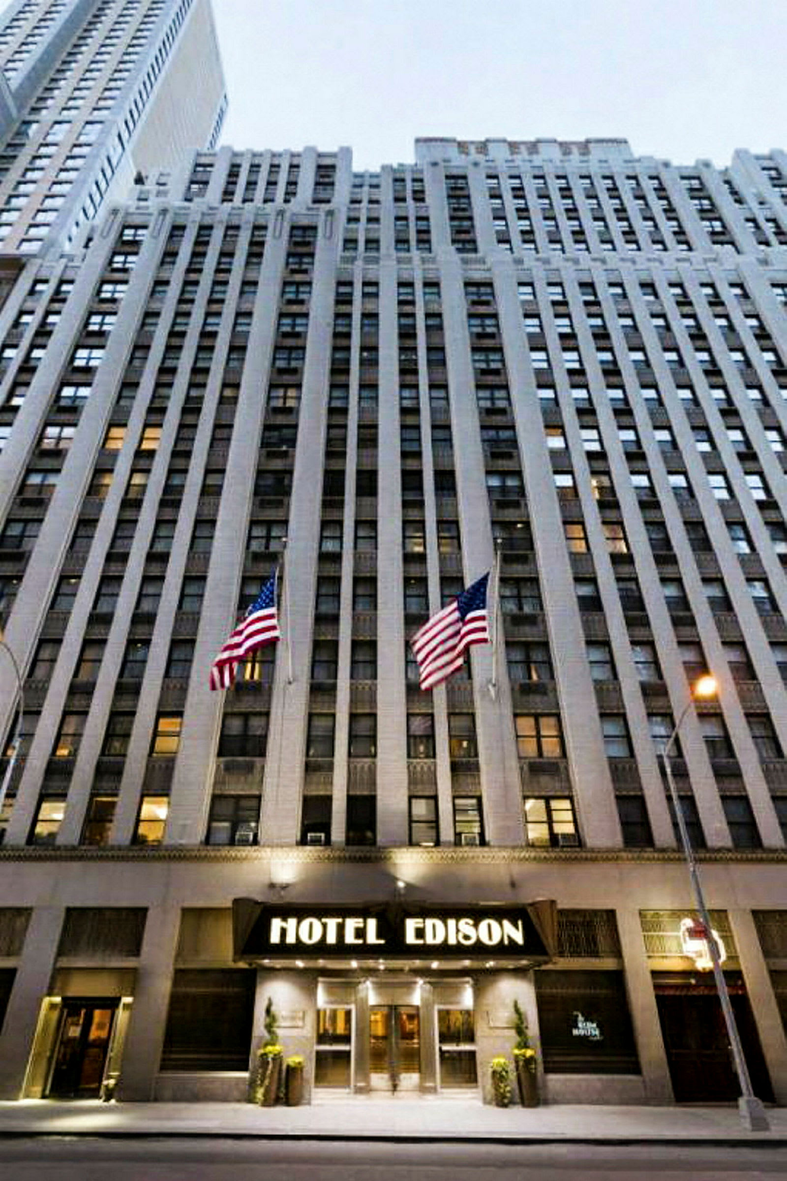 Näkymä korkeaan rakennukseen, jonka sisäänkäynnissä lukee "Hotel Edison", ja sen edessä on kaksi Yhdysvaltain lippua. (Tekoälyn luoma teksti)