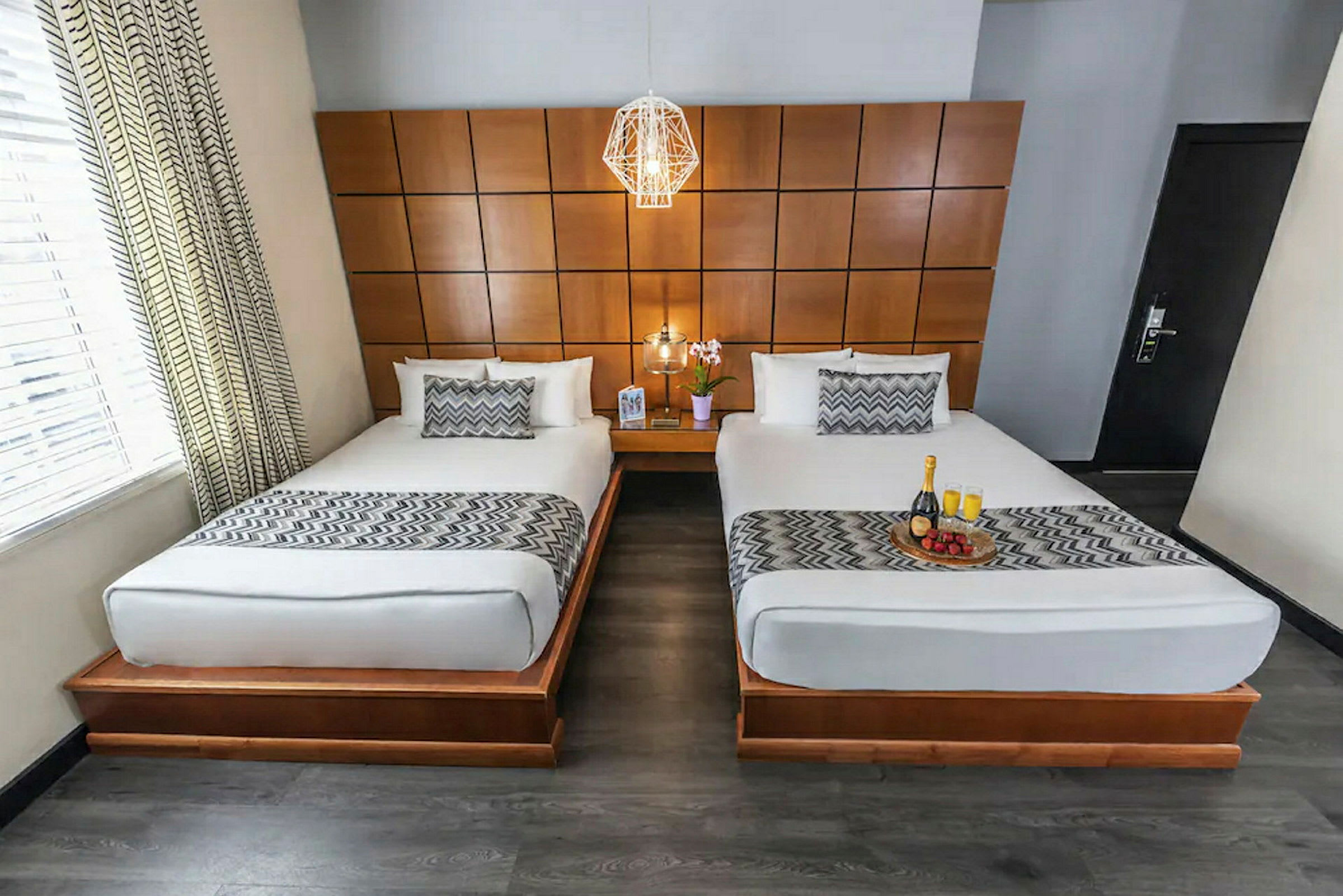 Kuva hotellihuoneesta, jossa on kaksi erillistä sänkyä valkoisilla vuodevaatteilla ja kuvioiduin päiväpeittein. Huoneessa on puupaneeliseinä ja sänkyjen välissä yöpöytä, jolla on kukka. (Tekoälyn luoma teksti)