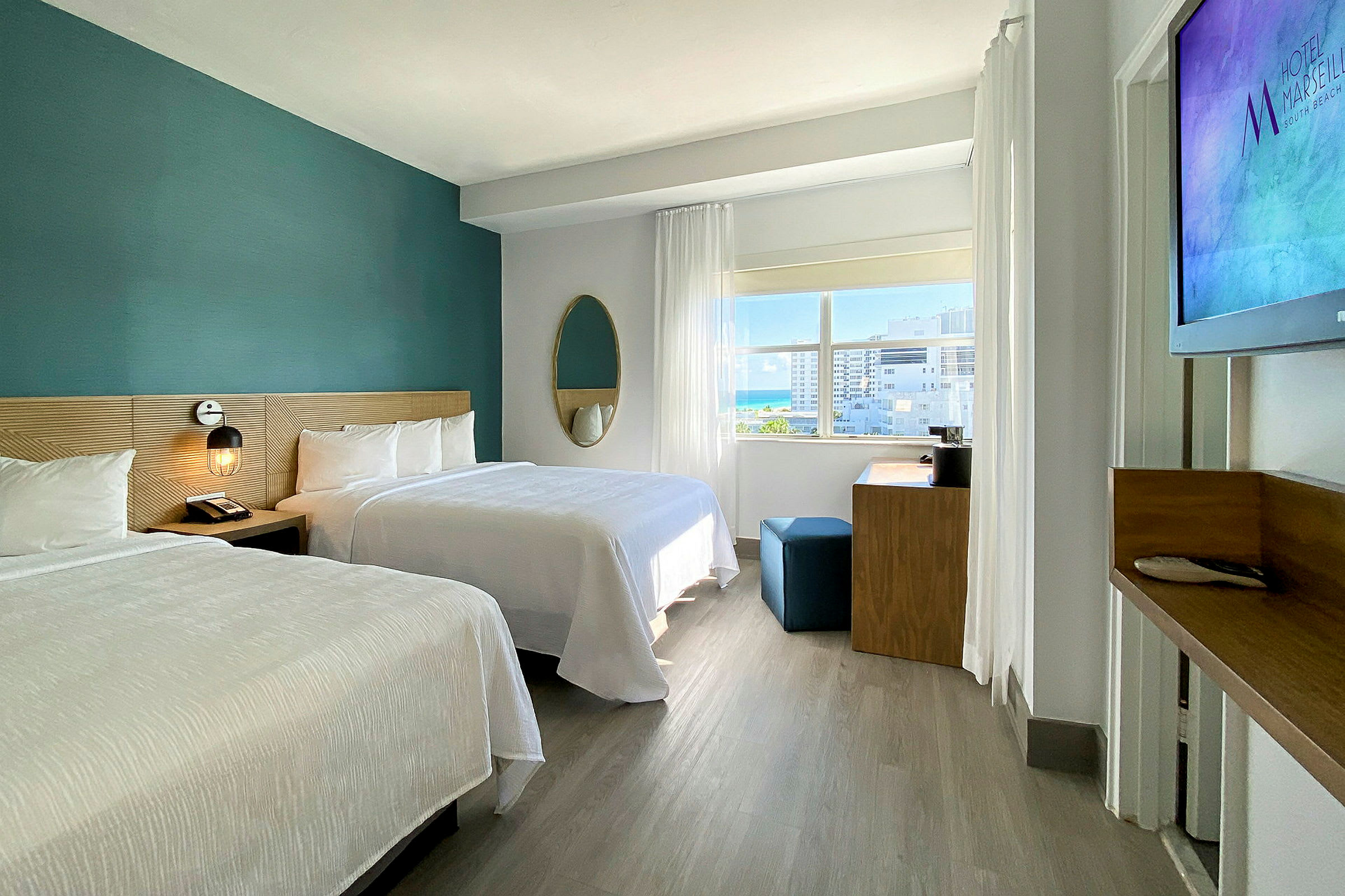 Moderni hotellihuone, jossa on kaksi parisänkyä, työpöytä ja suuri ikkuna, josta pääsee luonnonvaloa sisään. (Tekoälyn luoma teksti)