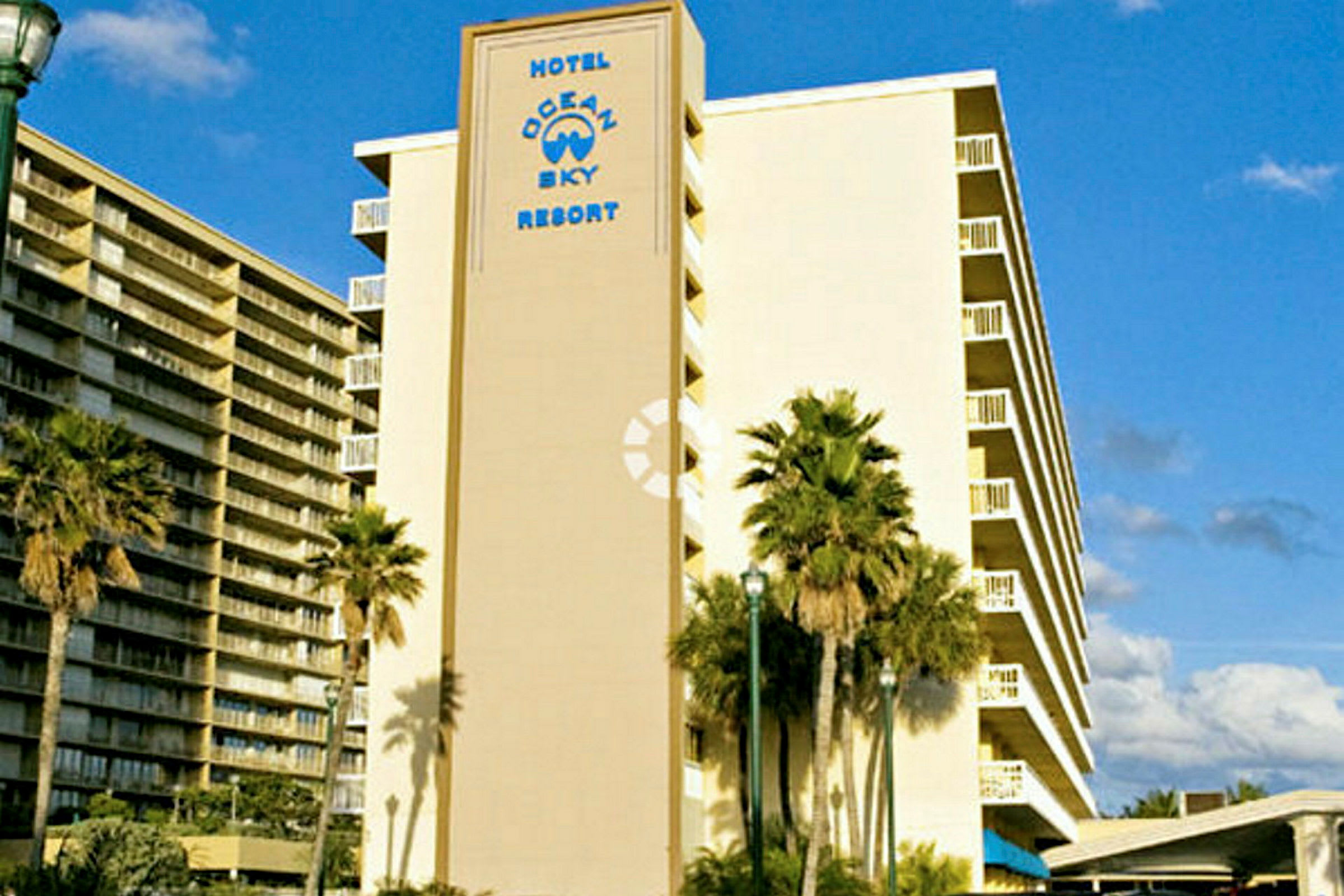 Kuvassa hotelli, jonka julkisivussa lukee "Hotel Ocean Sky Resort". Rakennuksen edessä näkyy palmupuita. (Tekoälyn luoma teksti)