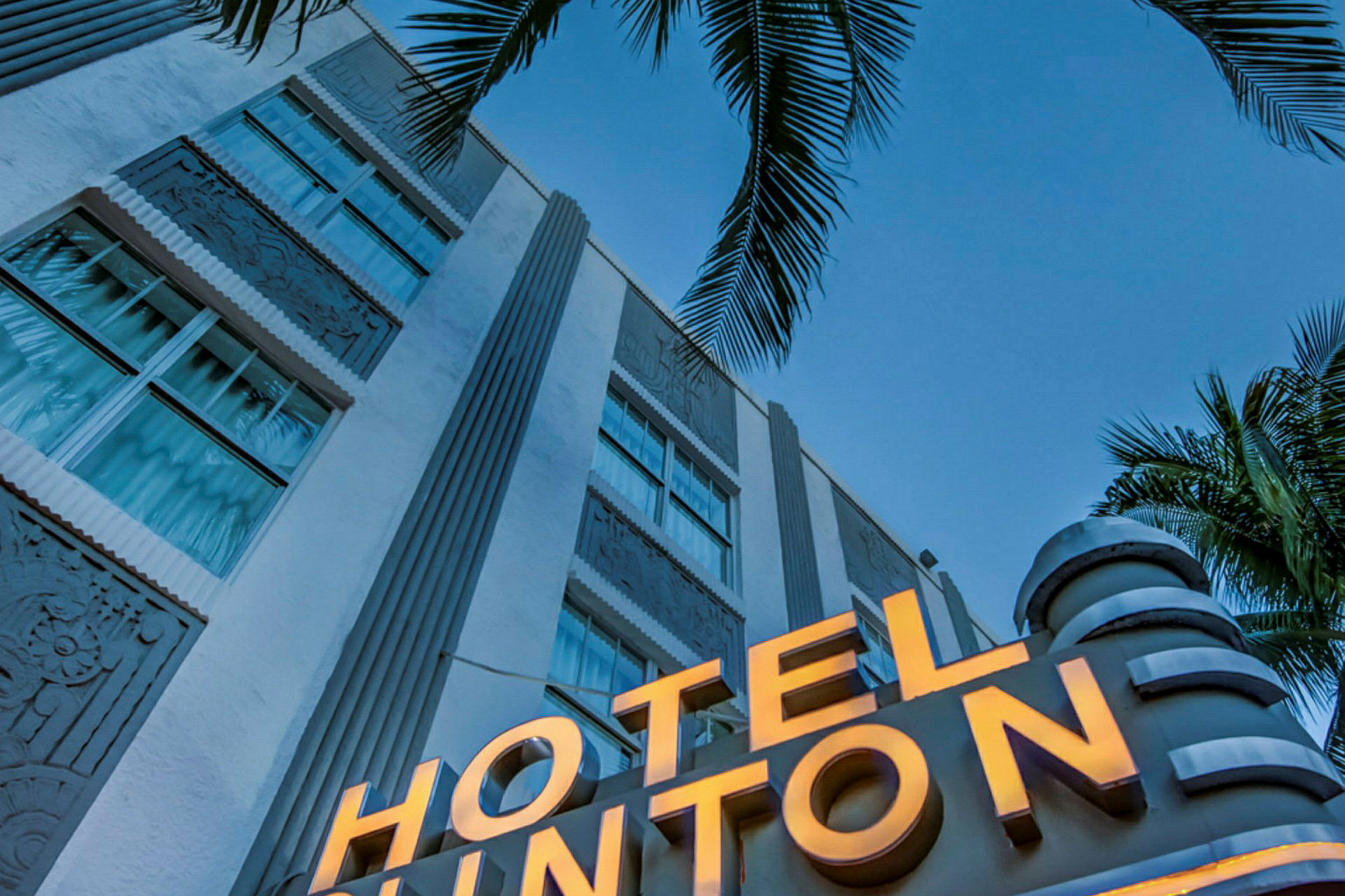 Lähikuva valkoisesta rakennuksesta, jossa valaistu kyltti lukee "HOTEL", ja etualalla on palmuja kirkasta sinistä taivasta vasten. (Tekoälyn luoma teksti)