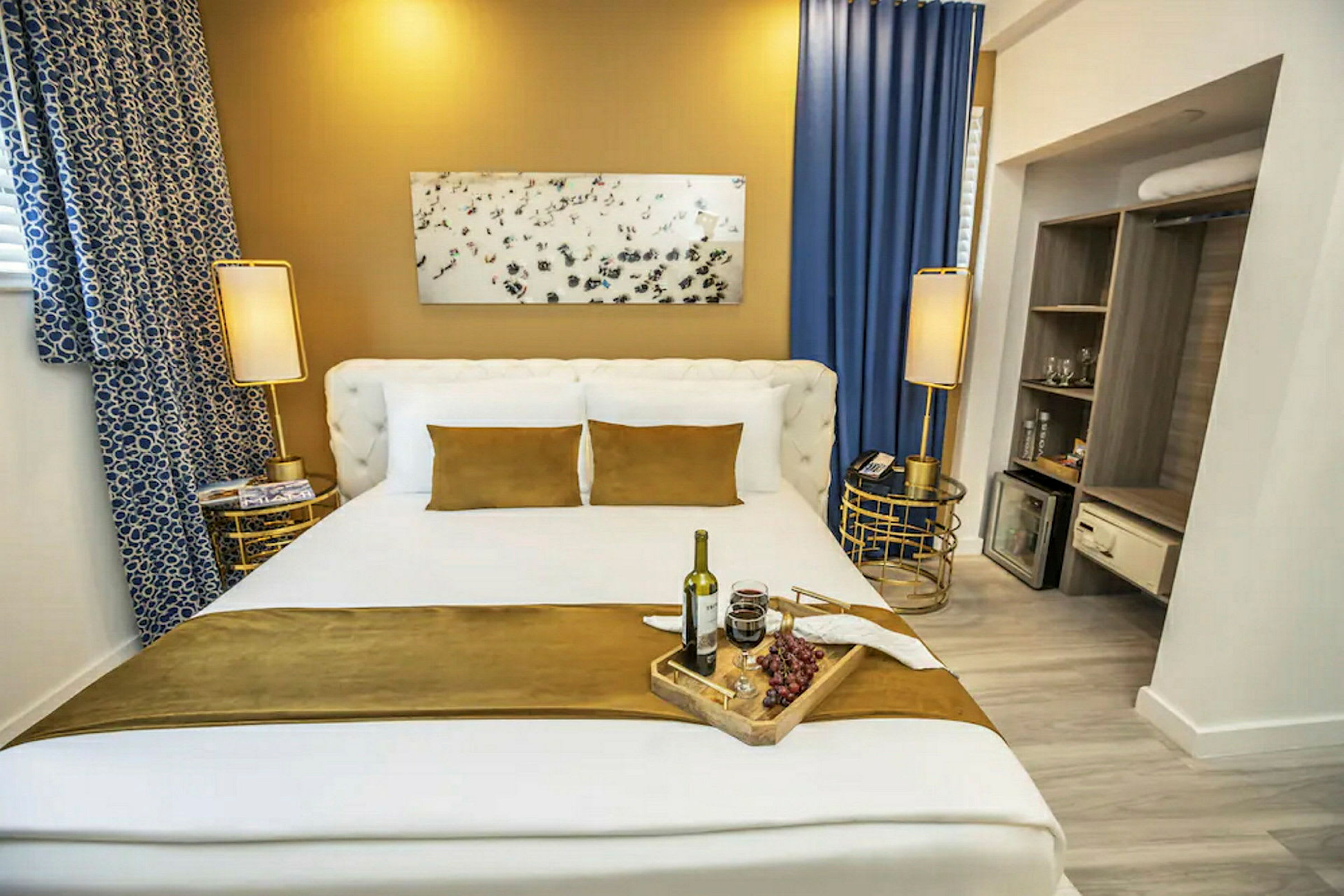 Hotellihuone, jossa on suuri sänky, vuodevaatteet valkoisina ja kullaruskean sävyisinä. Yöpöydät ja koristeet viimeistelevät huoneen. (Tekoälyn luoma teksti)