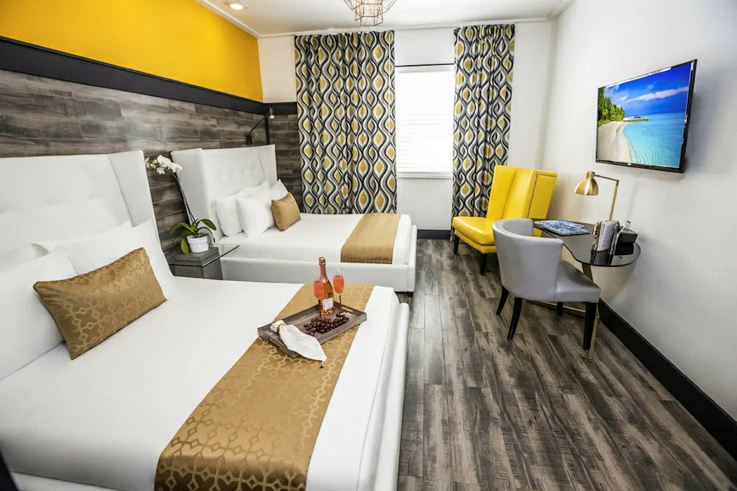 Hotellihuone, jossa on kaksi valkoista sänkyä, keltaisen ja harmaan sävyinen sisustus sekä seinällä televisio. (Tekoälyn luoma teksti)