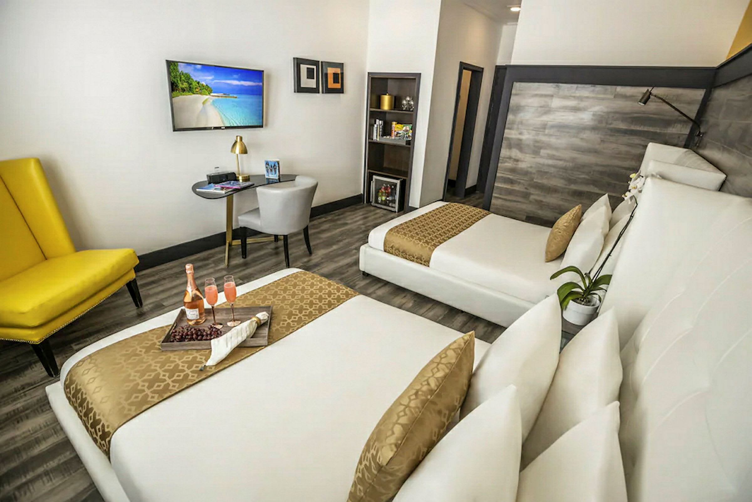 Moderni hotellihuone, jossa on kaksi sänkyä, keltainen nojatuoli, työpöytä ja seinällä taulutelevisio. (Tekoälyn luoma teksti)