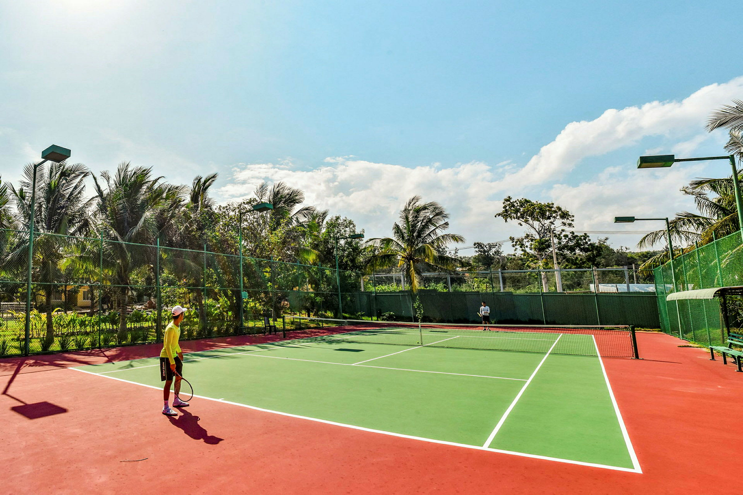 Kaksi henkilöä pelaa tennistä ulkokentällä, jota ympäröivät vehreys ja palmut kirkkaan taivaan alla. (Tekoälyn luoma teksti)
