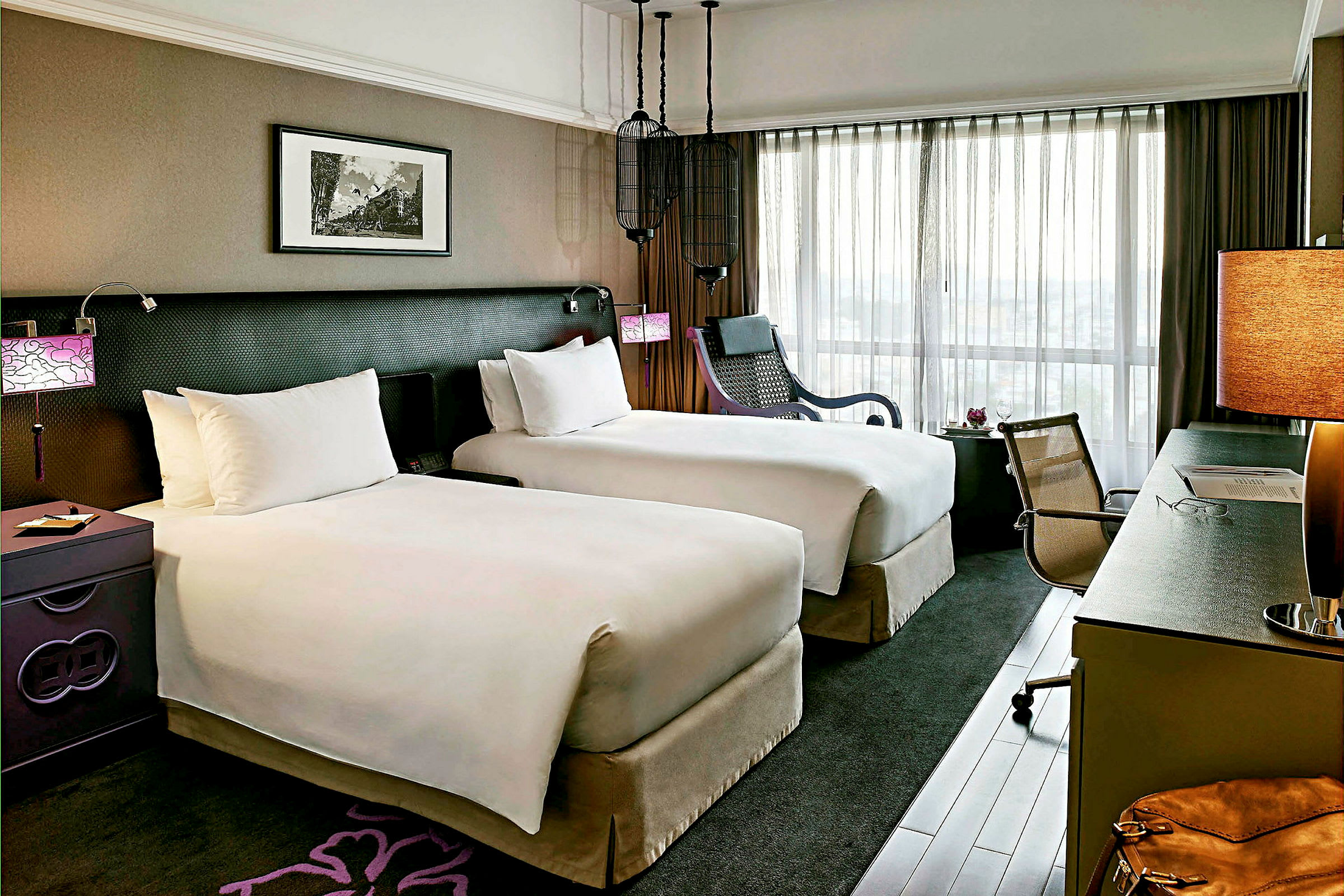 Hotellihuone, jossa kaksi yhden hengen vuodetta, ikkuna läpinäkyvillä verhoilla, kirjoituspöytä tuoleineen ja nojatuoli. (Tekoälyn luoma teksti)