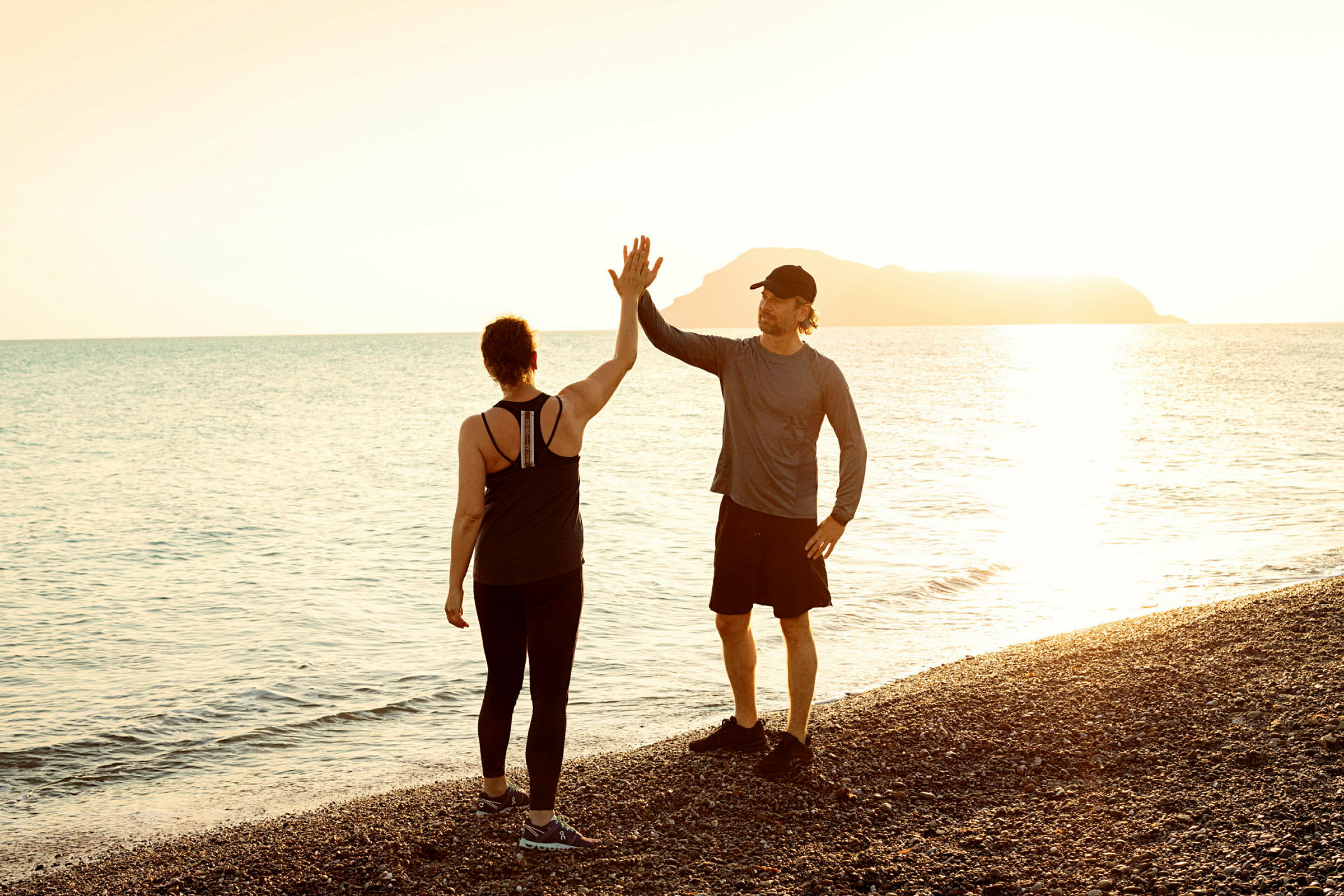 Kaksi henkilöä seisoo kivirannalla ja tekevät high five -tervehdyksen auringonlaskun aikana meren yllä. (Tekoälyn luoma teksti)