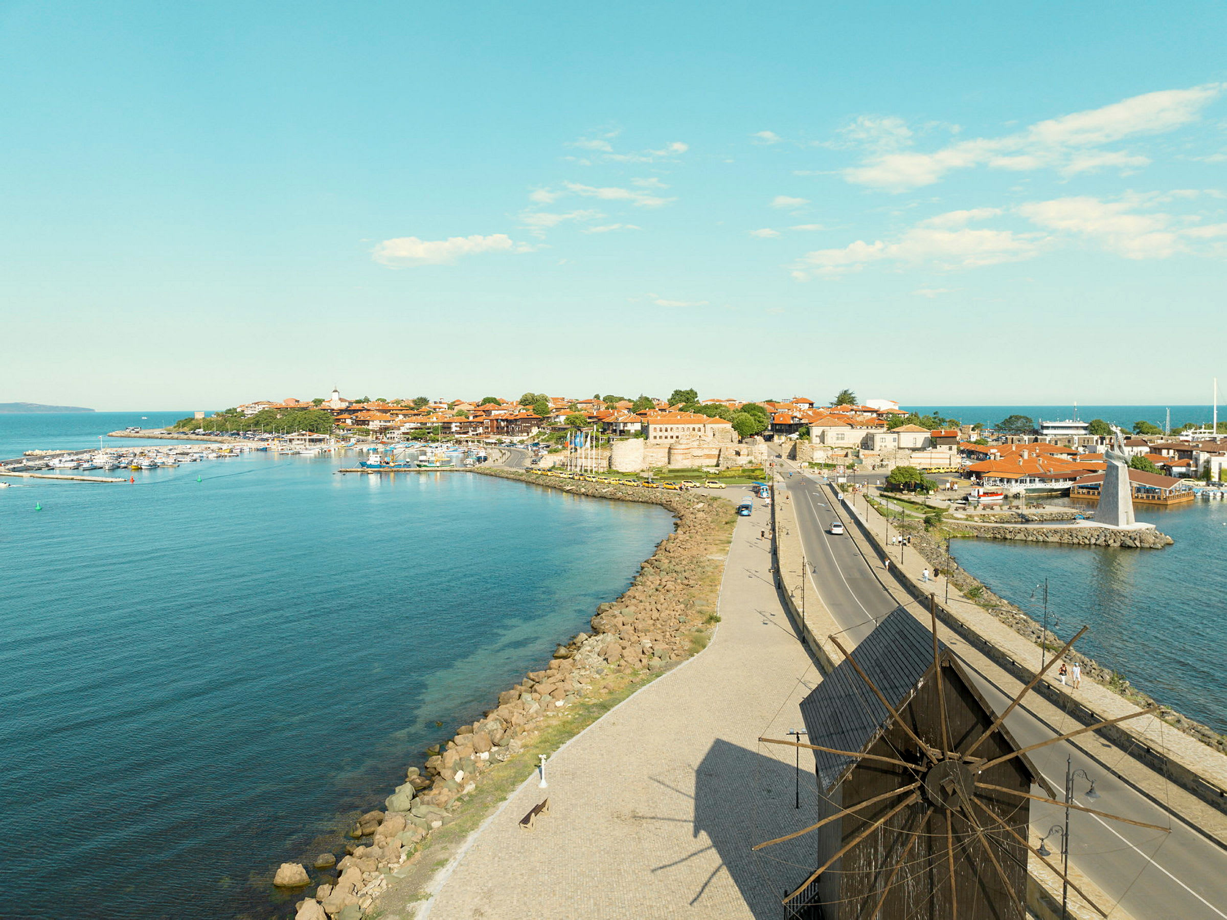 Rannikolla sijaitseva kaupunki, rakennuksia, mylly ja tie etualalla. (Tekoälyn luoma teksti)