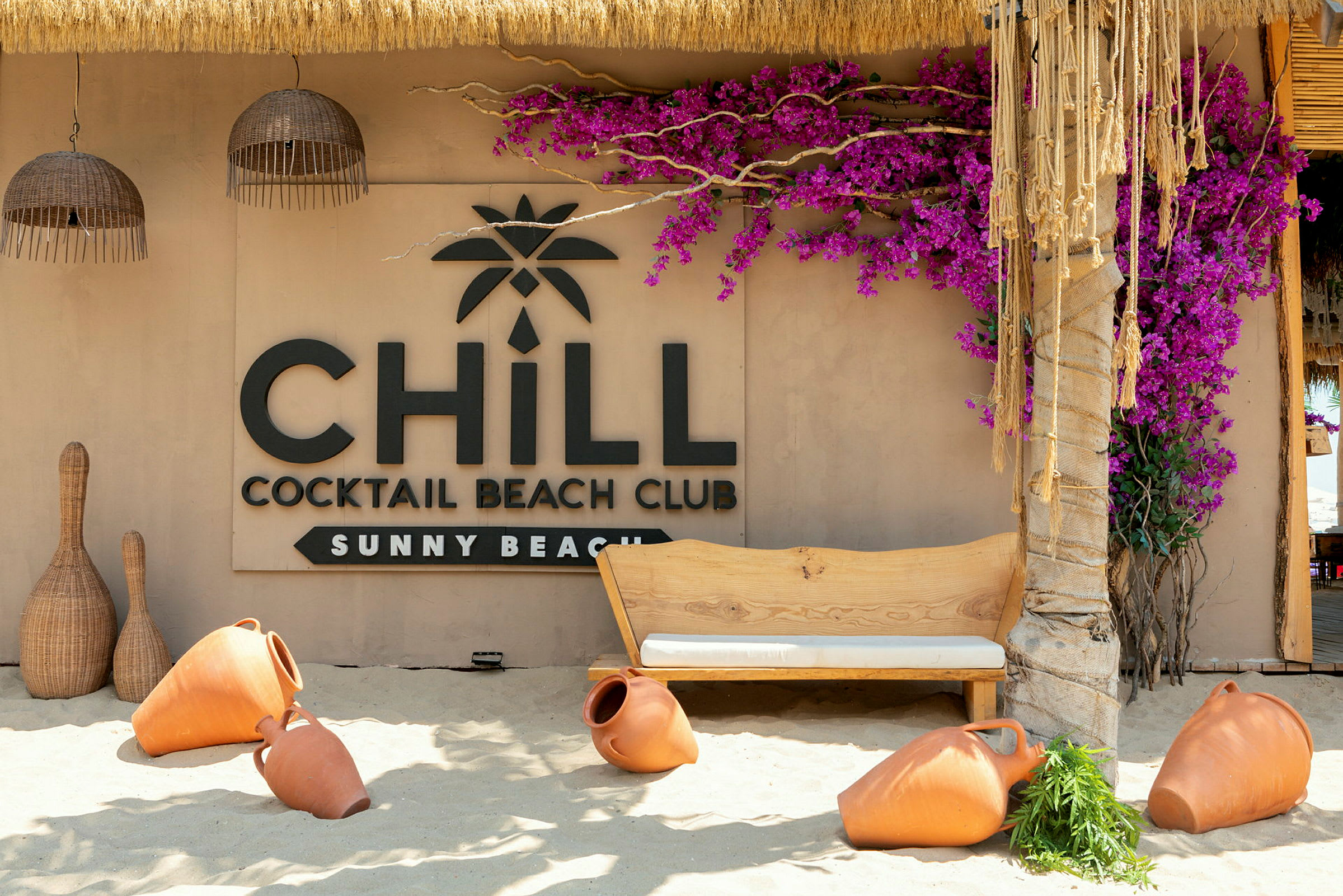Rantabaari, jossa kyltti 'Chill Cocktail Beach Club Sunny Beach', ympäröitynä terrakottaruukuilla ja värikkäillä kukilla. (Tekoälyn luoma teksti)