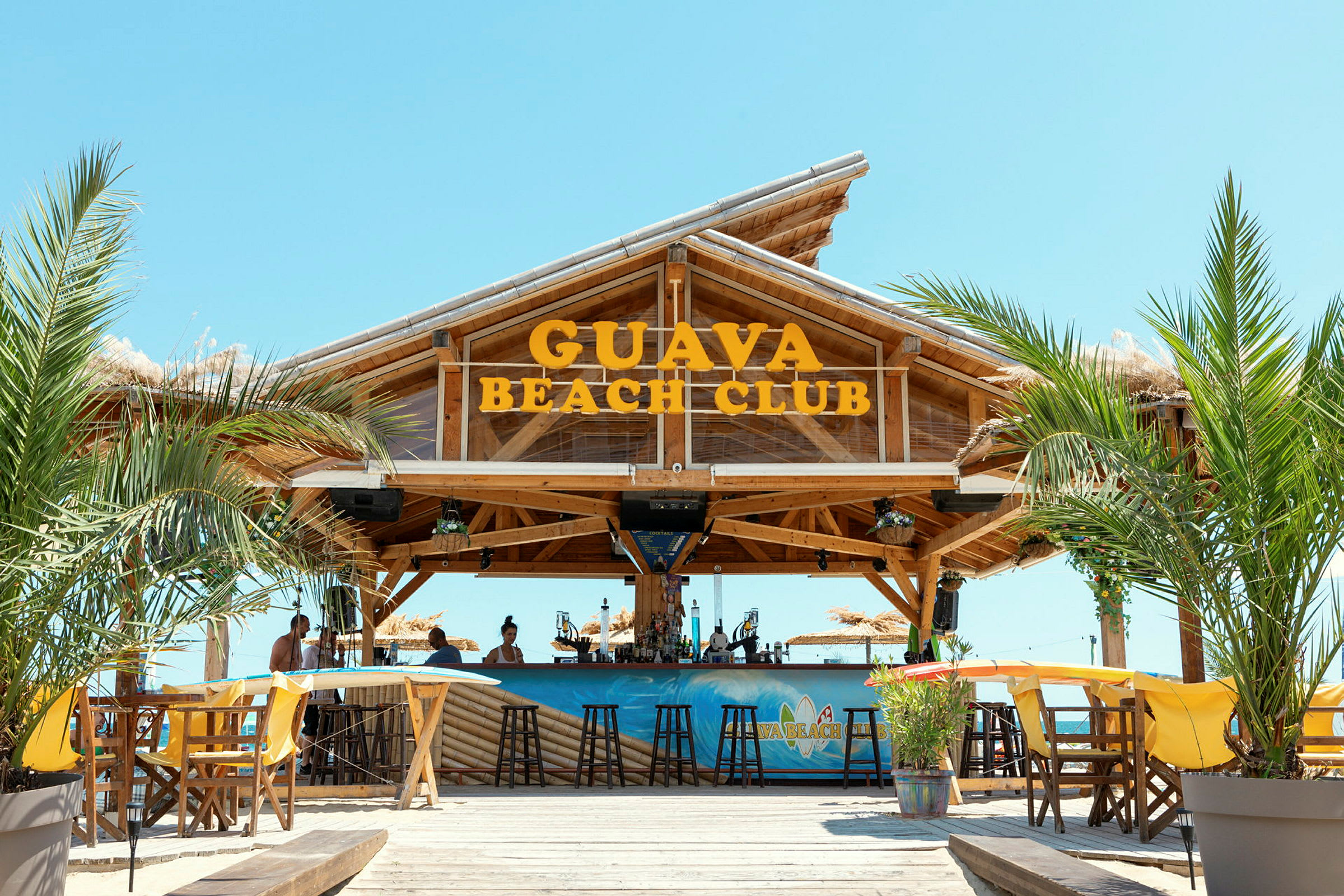 Kuva Guava Beach Clubista, puurakenteisesta baarista, istuma-alueista, palmupuista ja kirkkaasta taivaasta. (Tekoälyn luoma teksti)