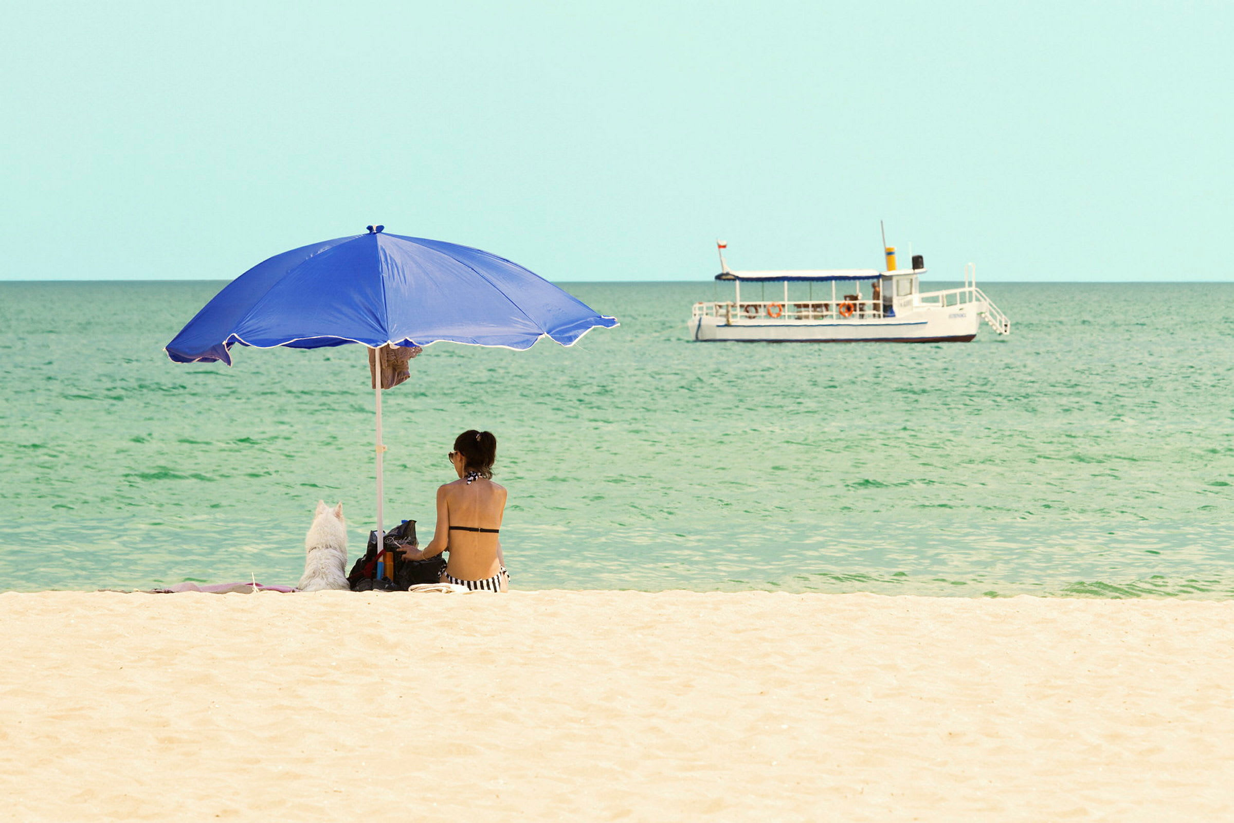 Henkilö istuu sinisen aurinkovarjon alla hiekkarannalla, taustalla meri. Valkoinen vene näkyy vedessä. (Tekoälyn luoma teksti)